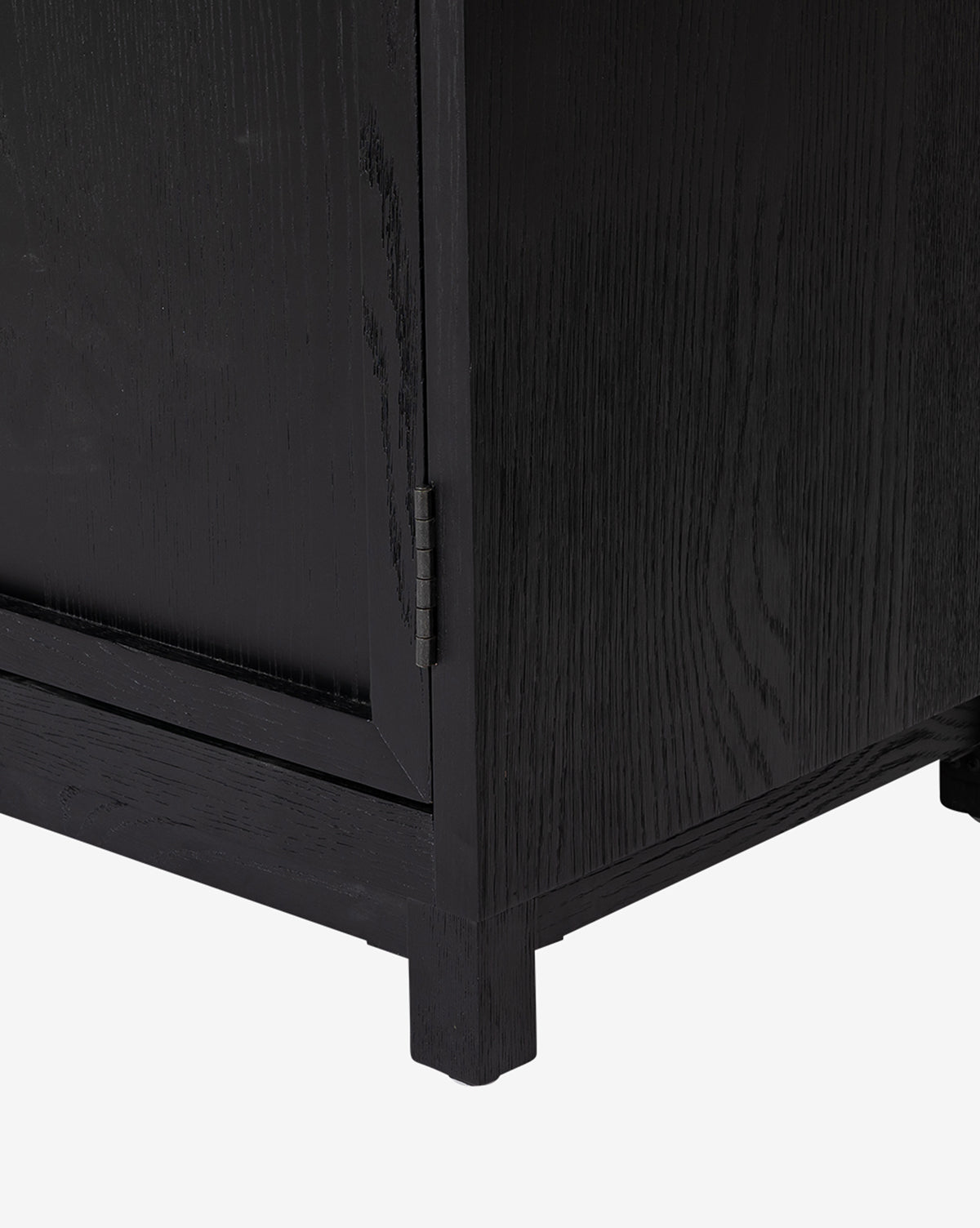 Close-up of the Emrys Cabinet’s bottom corner reveals detailed wood grain, sturdy short square legs, and a cabinet door hinge, showcasing its elegant shelving design against a white background, by McGee & Co.