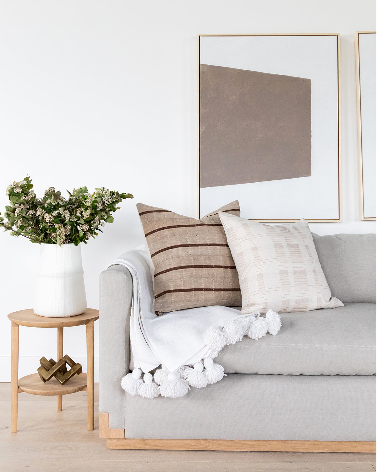 A light gray sofa with neutral pillows and a white tasseled throw sits by a wooden side table topped with a white vase and greenery, set against a white wall decorated with curated items and the Entering Shape 1 taupe wall art.