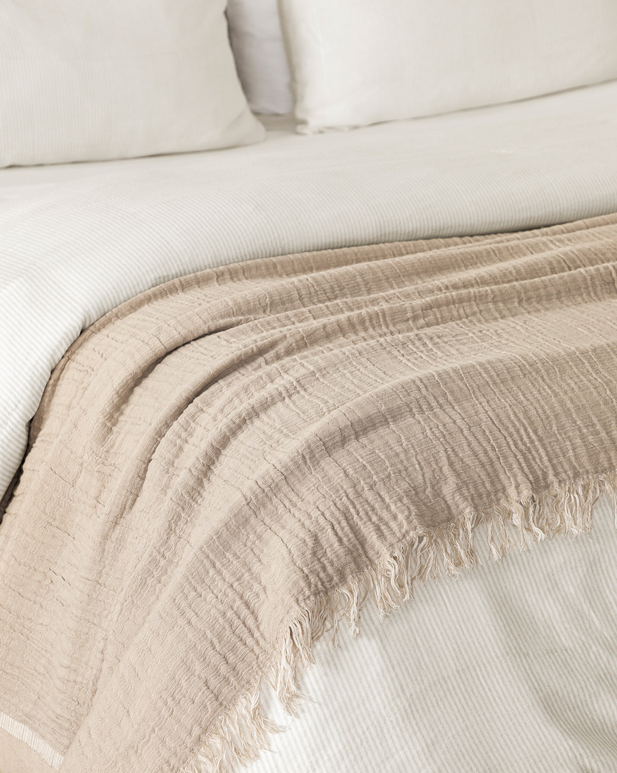 The Ermessa Oversized Throw, a beige textured blanket with fringed edges made from a soft cotton-bamboo blend, is draped over a neatly made bed with white bedding and pillows.