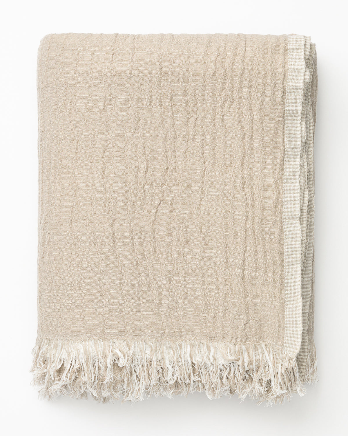 The Ermessa Oversized Throw, made from a soft cotton-bamboo blend, features textured beige fabric with fringed edges and a woven border, and is shown neatly folded on a white background.