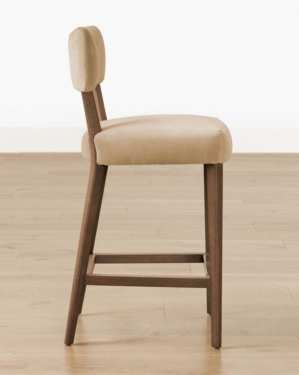 Certainly! Here’s your blended description with the exact string at the end: Side view of the Makerspalm Etienne Stool (Ready to Ship), featuring a light beige cushioned seat and wooden angled legs, displayed on a light wood floor with a plain white wall background. McGee & Co.