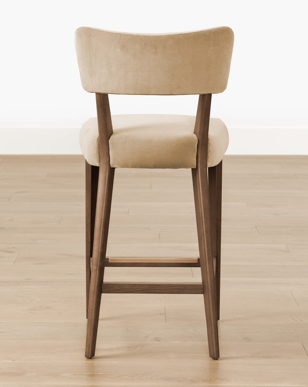 The Etienne Stool (Ready to Ship) by Makerspalm features beige upholstery, a curved backrest, and wooden legs, shown from the back on a light wood floor with a white wall background. McGee & Co.