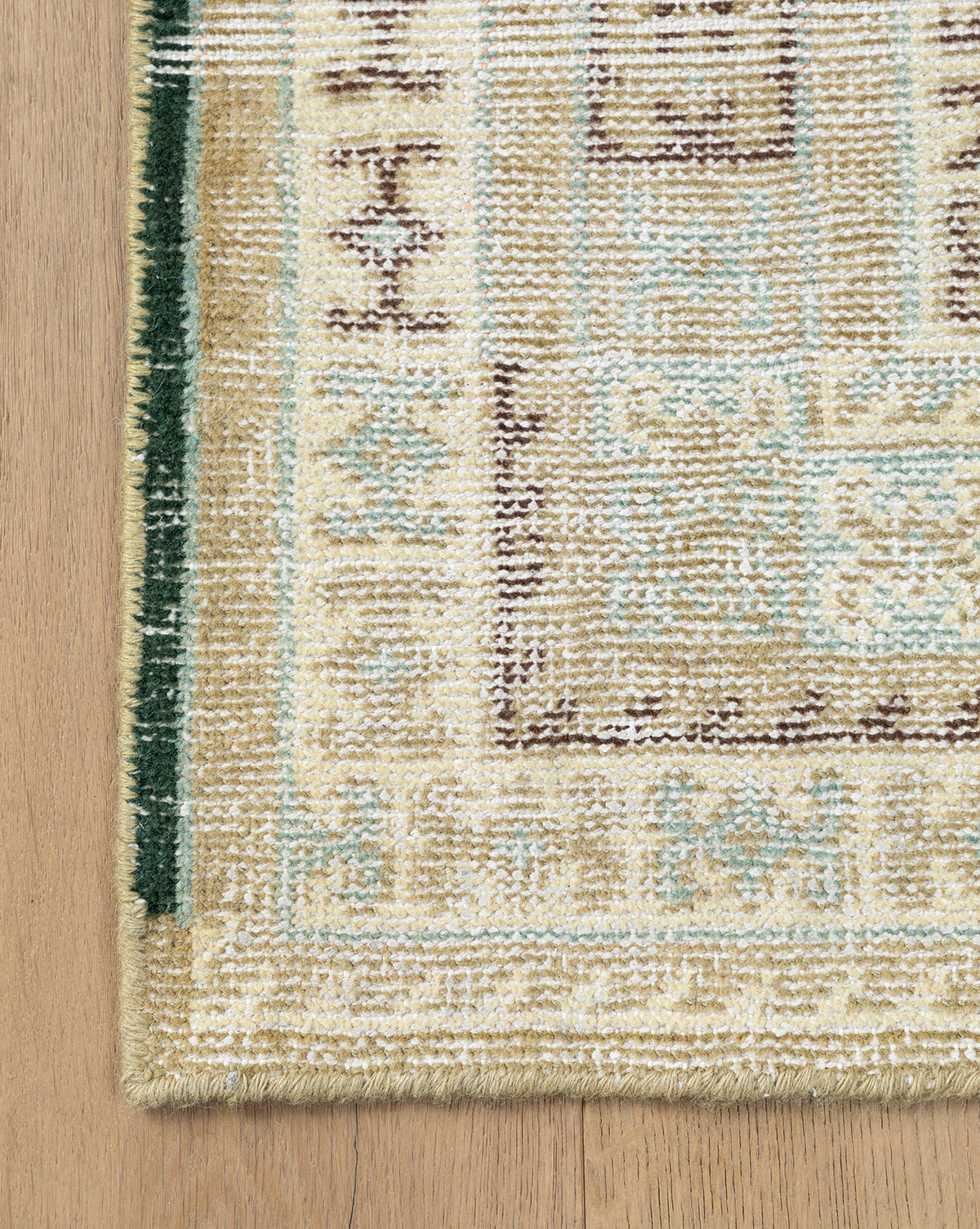 Corner of the Evangeline Hand-Knotted Wool Rug, shown in cream, beige, and pale blue on a wooden floor. This durable wool rug features a green border with intricate geometric patterns, by McGee & Co.