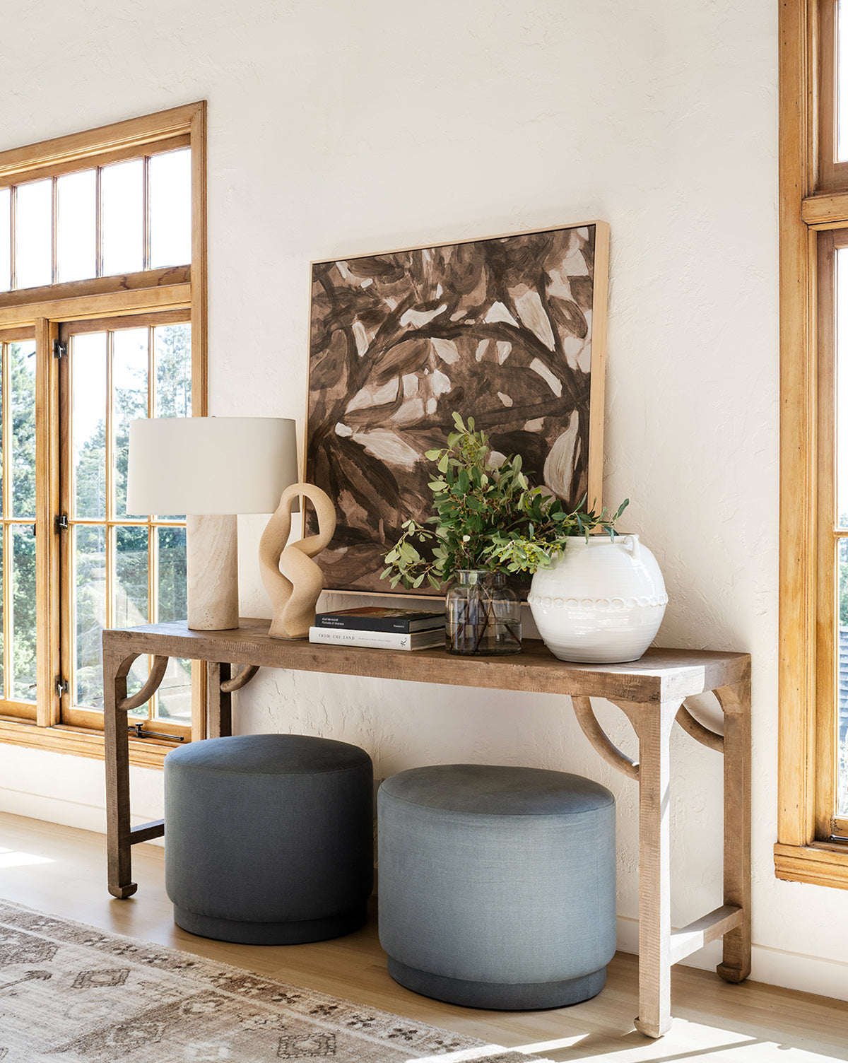 A wooden console table against a white wall holds a lamp, sculpture, books, leafy plant in a white vase, and the framed giclee canvas print Light of Summer by Cheryl Clinton; two blue ottomans are tucked beneath, with windows on either side.