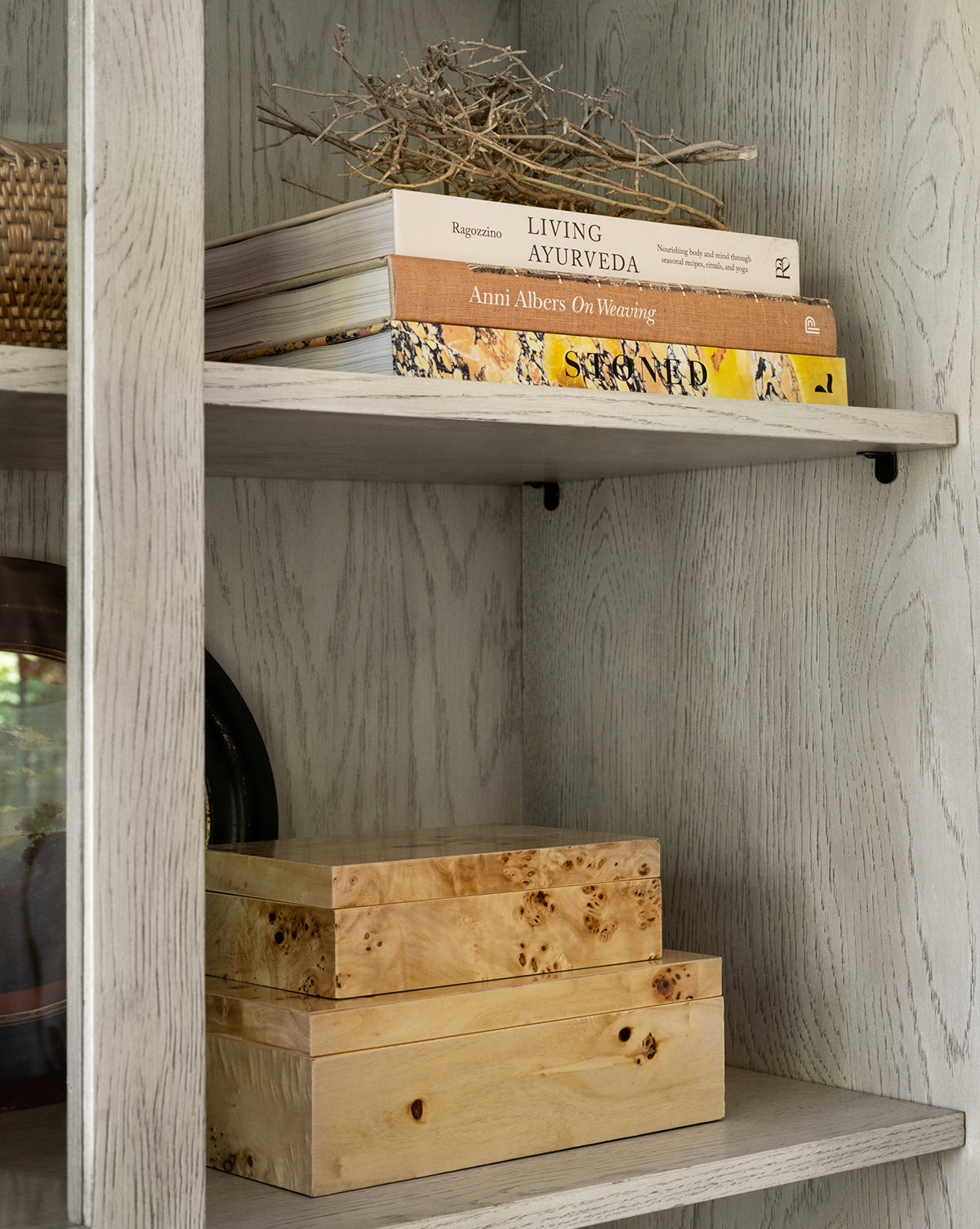 A bookshelf with light wood shelves displays a bundle of twigs, three stacked books, the Burl Wood Box, another wooden box, and a woven basket for a natural yet contemporary minimalist look.