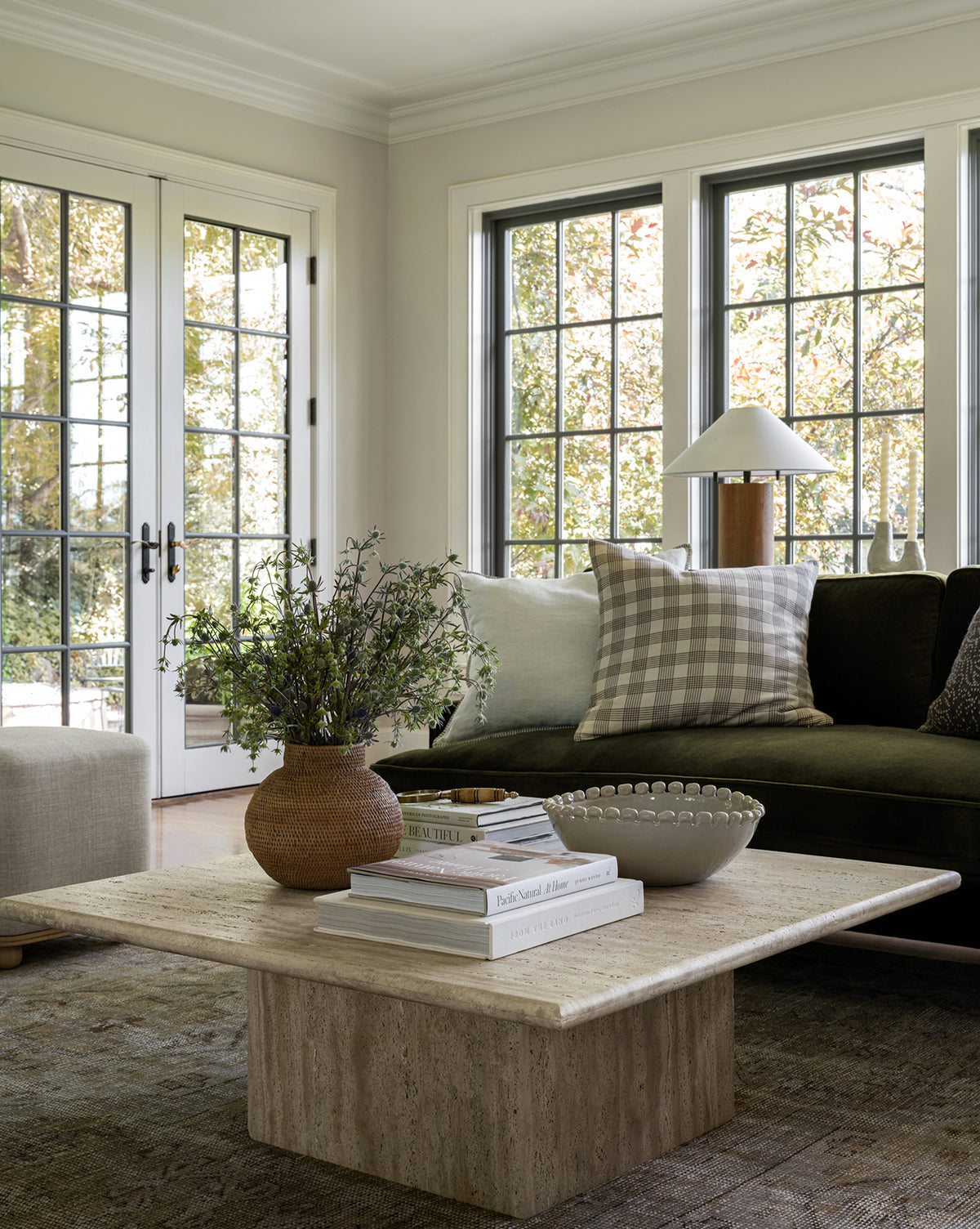 A bright living room with large windows, a dark sofa accented by light and checkered pillows, a square stone coffee table holding books, greenery in a vase, and the Malibu Bowl as décor. Trees are visible outside, by McGee & Co.