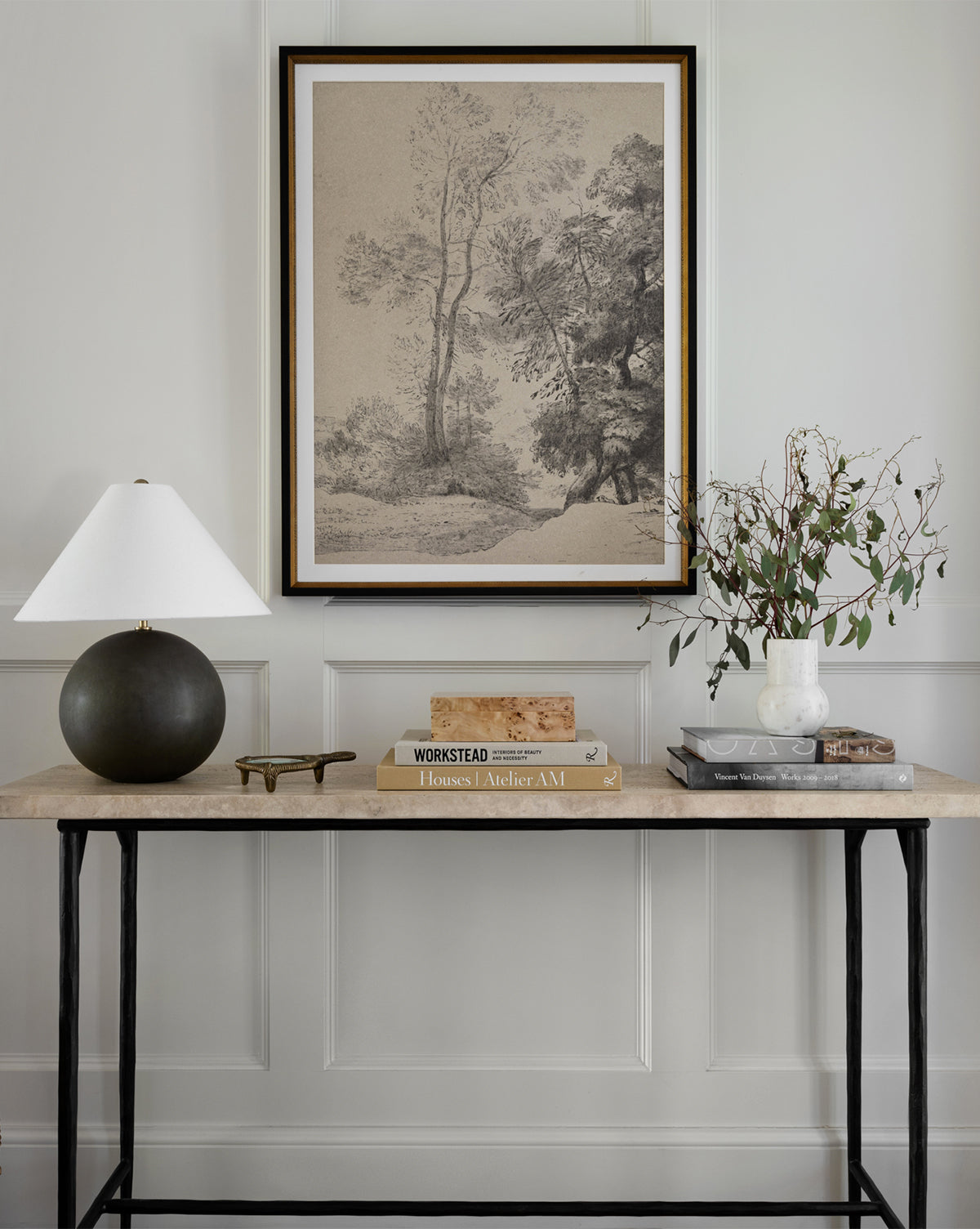 The console table holds a round black lamp, books, a white vase with leafy branches, and glasses against a white paneled wall beneath Pastoral Sketch, vintage landscape art in a distressed wood frame.