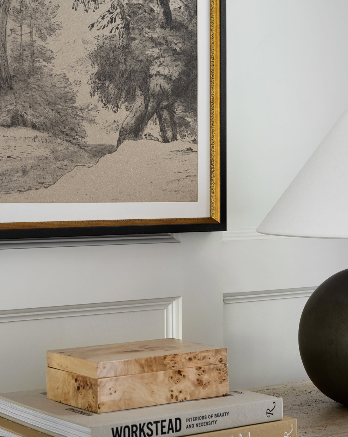 A close-up of a wall featuring the Pastoral Sketch rustic wall decor—a distressed wood frame with vintage landscape art—beside a table holding books, a wooden box, and a lamp with a white shade and dark base.