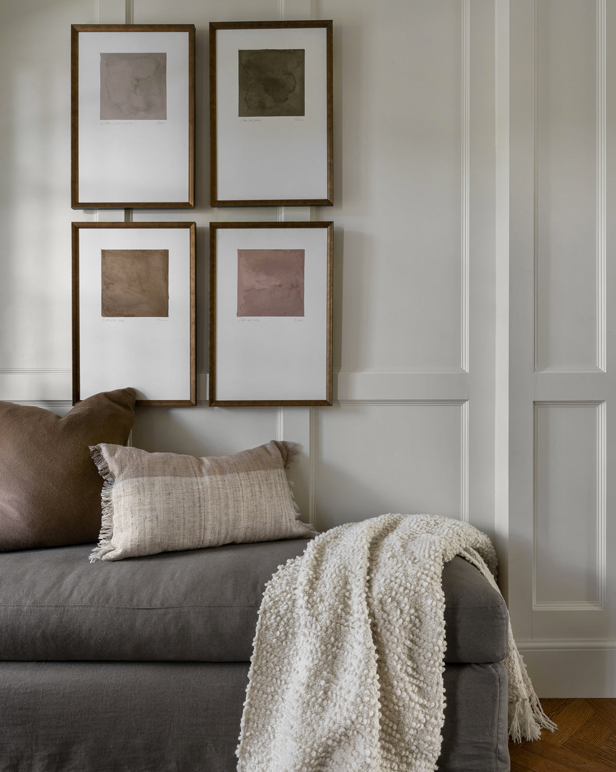 A cozy gray sofa with a beige pillow and cream throw sits against a white paneled wall, topped by four gold-framed Fossil Study artworks featuring neutral abstract designs.