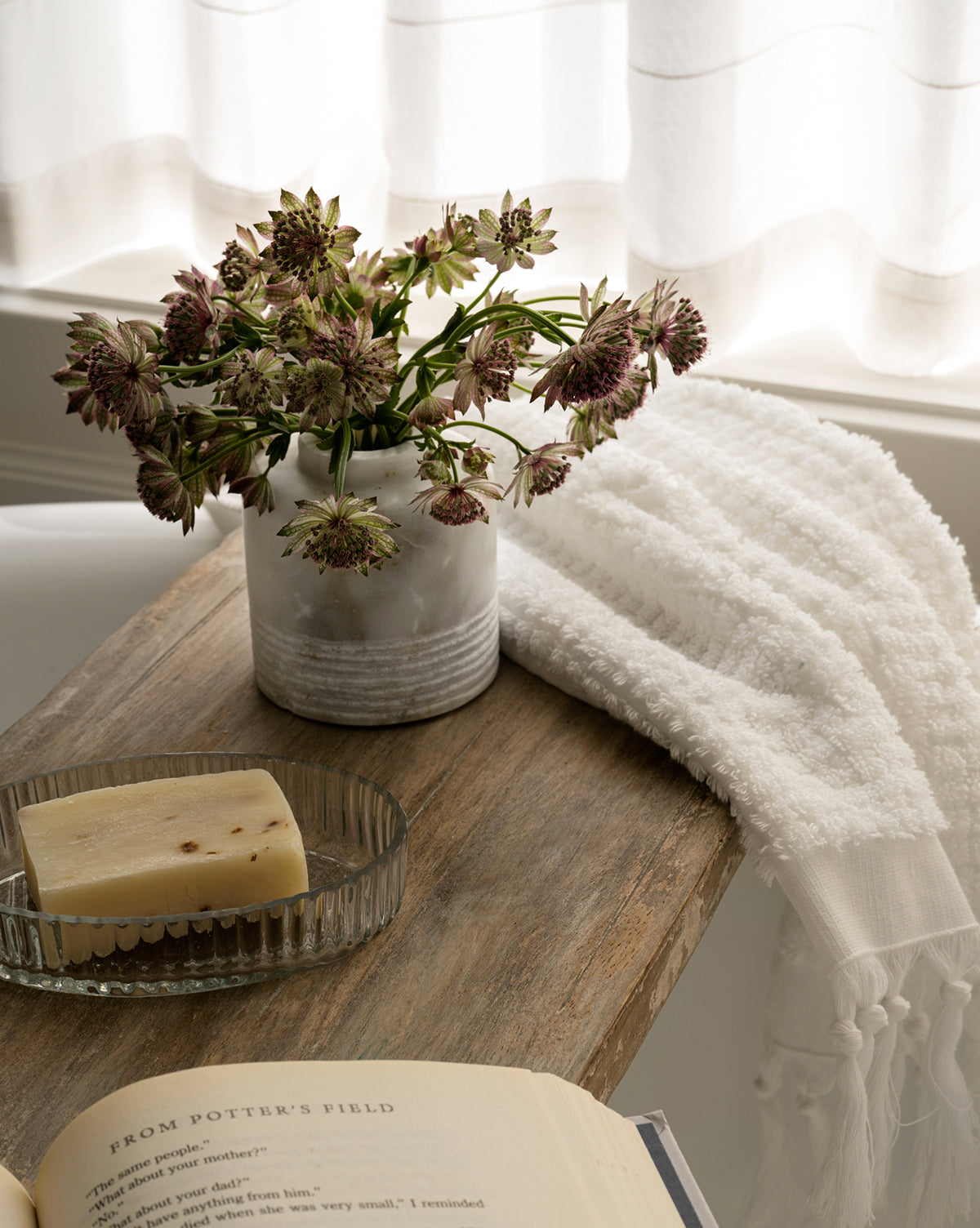 A small vase of flowers, a glass dish with soap, and an open book sit on the Rounded Edge Bath Caddy beside a white towel, while soft light filters through sheer curtains in the background.