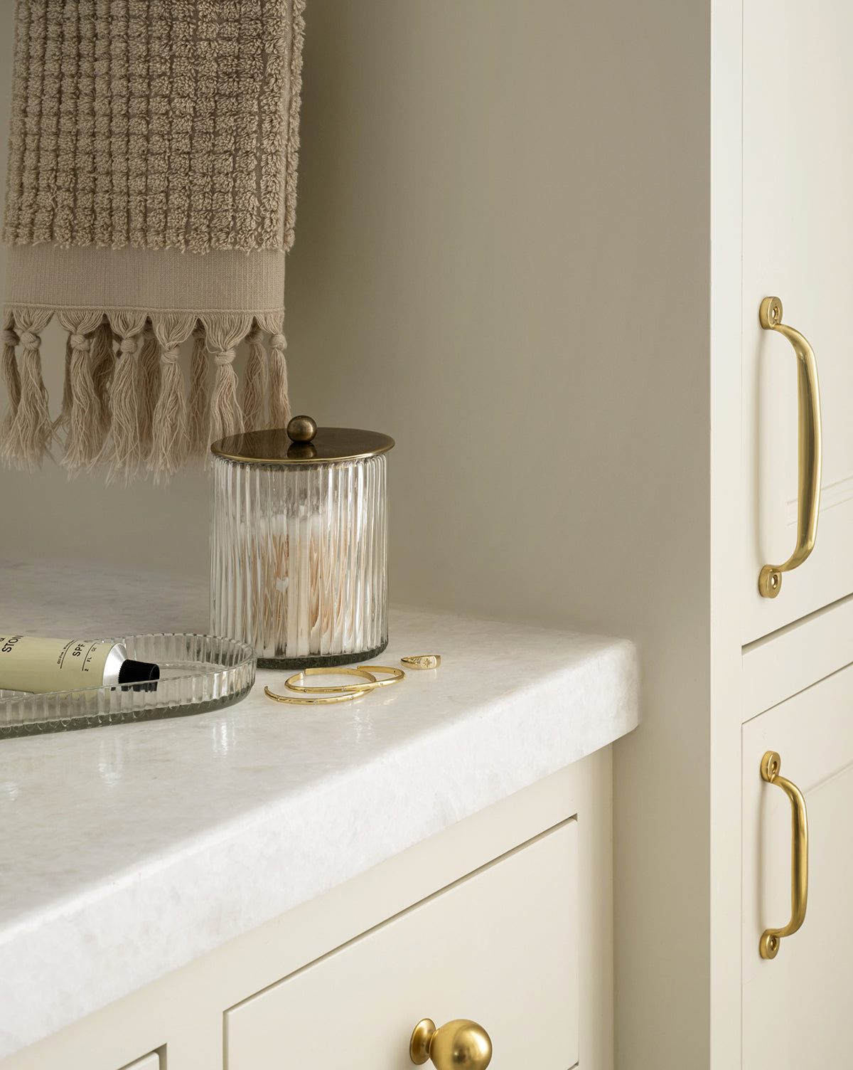 A cream bathroom countertop displays a textured towel, a glass jar from the Stein Glass Collection with brass lid, a glass tray holding a tube and gold bracelet, plus gold cabinet handles for an elegant, minimal look.