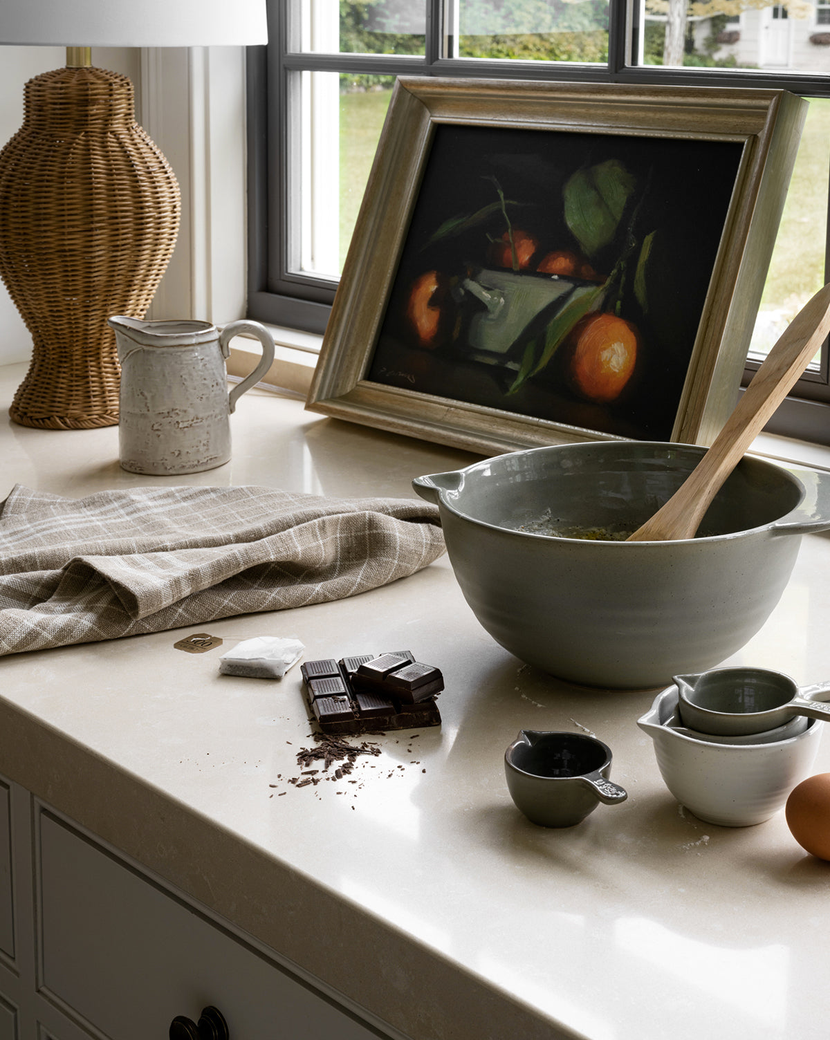 A kitchen counter with a mixing bowl, wooden spoon, measuring cups, a broken chocolate bar, an egg, a dish towel, a White Stoneware Pitcher, and a framed still-life painting beside a window by McGee & Co.