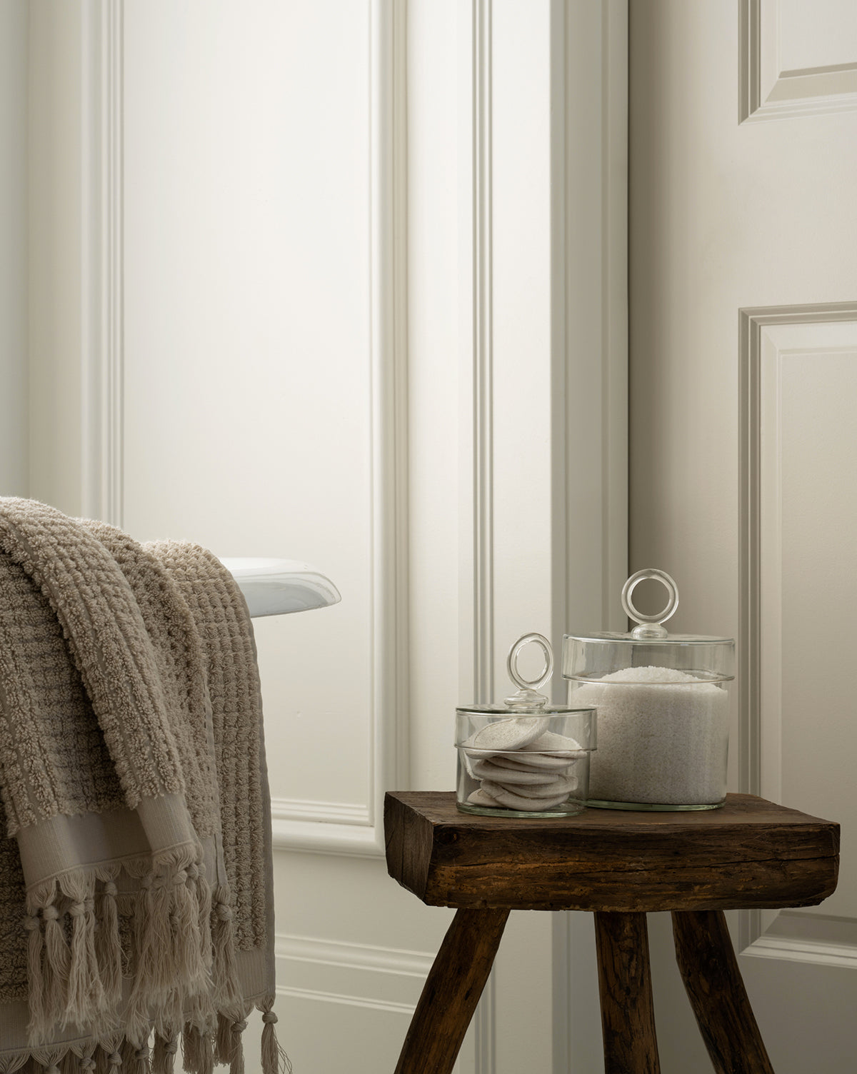 A cozy bathroom features a beige textured towel on the tub and a small wooden stool displaying Glass Lidded Canisters, ideal for storing cotton pads and bath salts, all set against white paneled walls by the countertop.