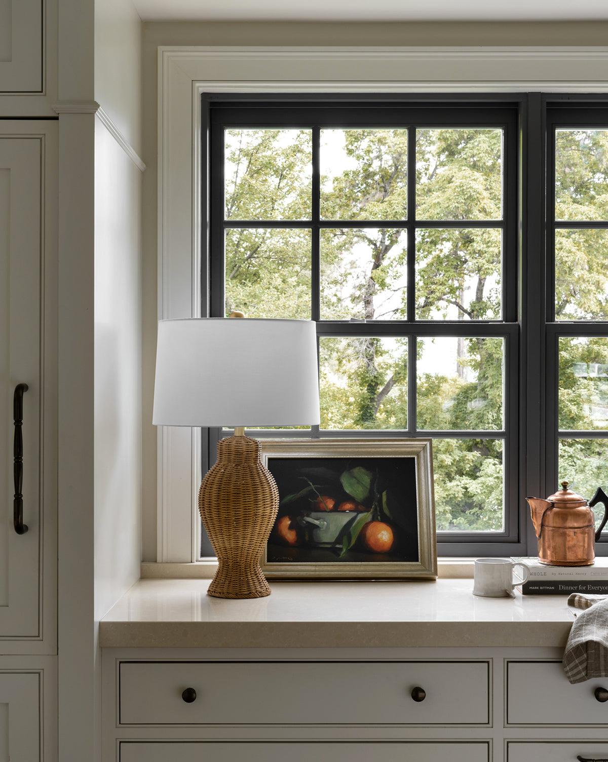 A kitchen countertop beneath a large window holds a wicker lamp with a white shade, the Oranges Still Life framed giclee print, a copper teapot, stacked books, and a light dish towel. Trees are visible outside.