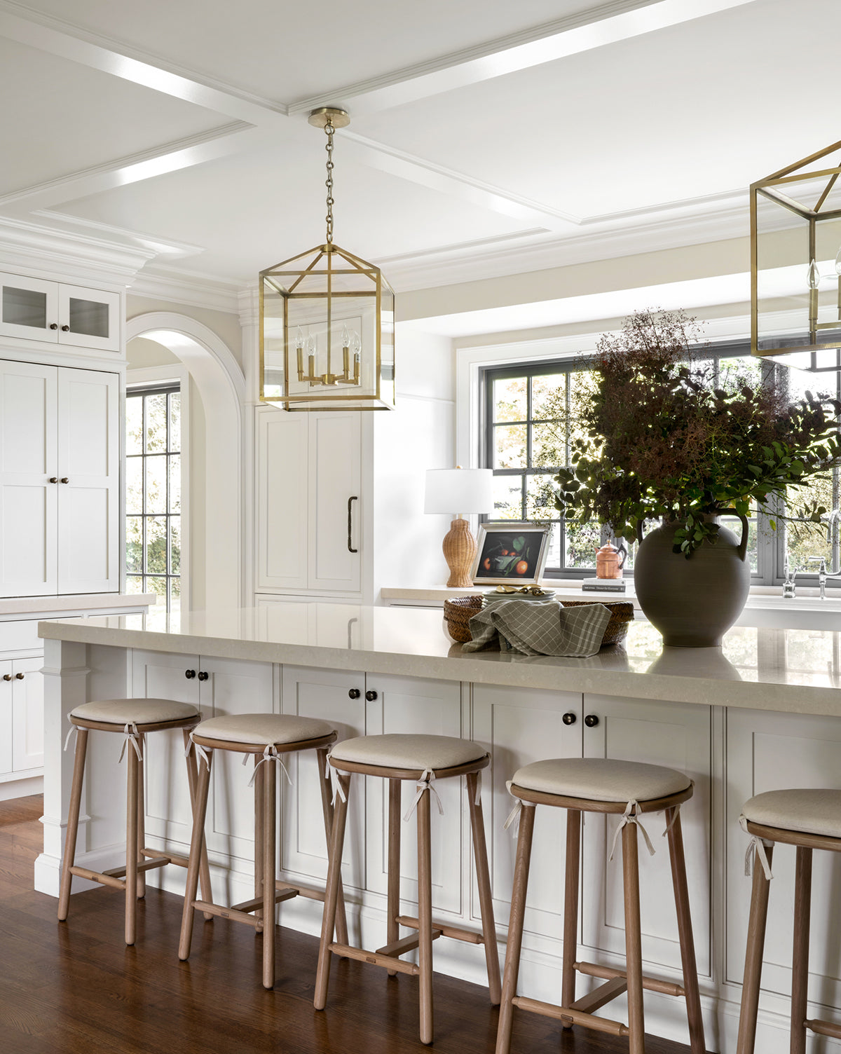 A bright, elegant kitchen with white cabinets, a spacious island with four Ralph Oak Counter Stools featuring linen cushions, gold pendant lights, a floral vase on the island, and a window above the sink streaming in natural light by McGee & Co.