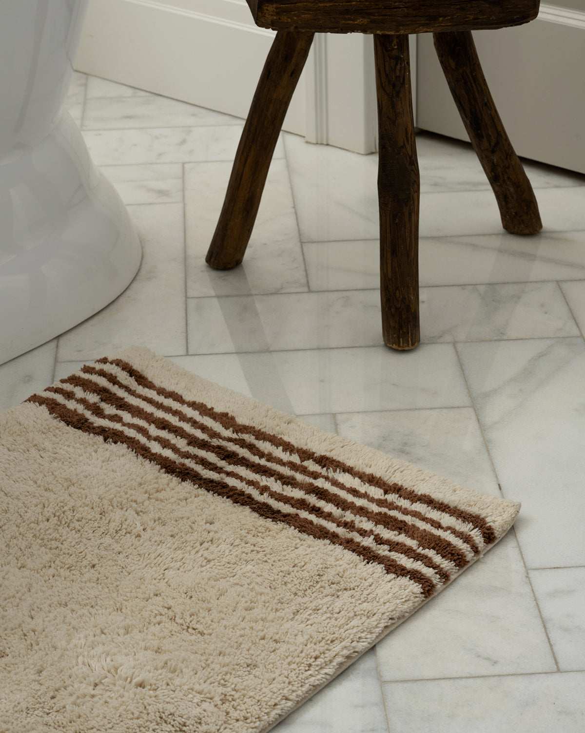 The Beata Bath Mat, featuring beige and brown stripes on plush cotton, lies on a white marble floor beside a rustic wooden stool and a white bathtub.