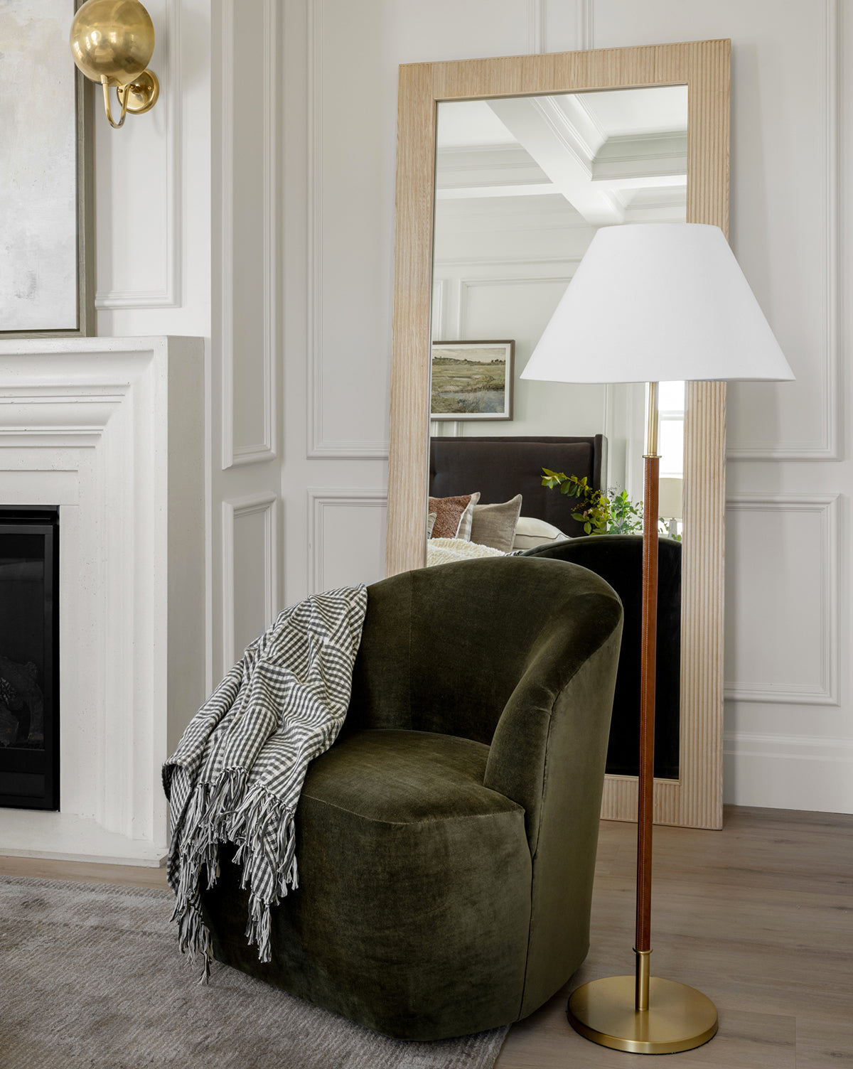 A cozy green velvet chair with a striped throw sits beside the Elijah Floor Lamp, featuring an elegant brass pole, next to a large wooden-framed mirror that reflects a stylish, modern living room with white paneled walls, by McGee & Co.
