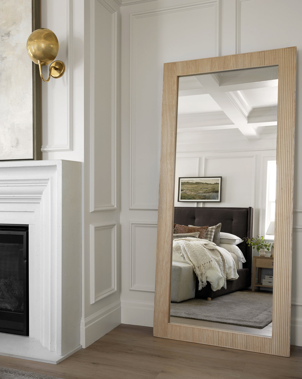A Rowan Floor Mirror with a ribbed oak frame leans against a white-paneled wall, reflecting a cozy bedroom scene with bed, pillows, nightstand, blanket, and landscape art. A gold wall sconce and part of a fireplace are also in view, by McGee & Co.