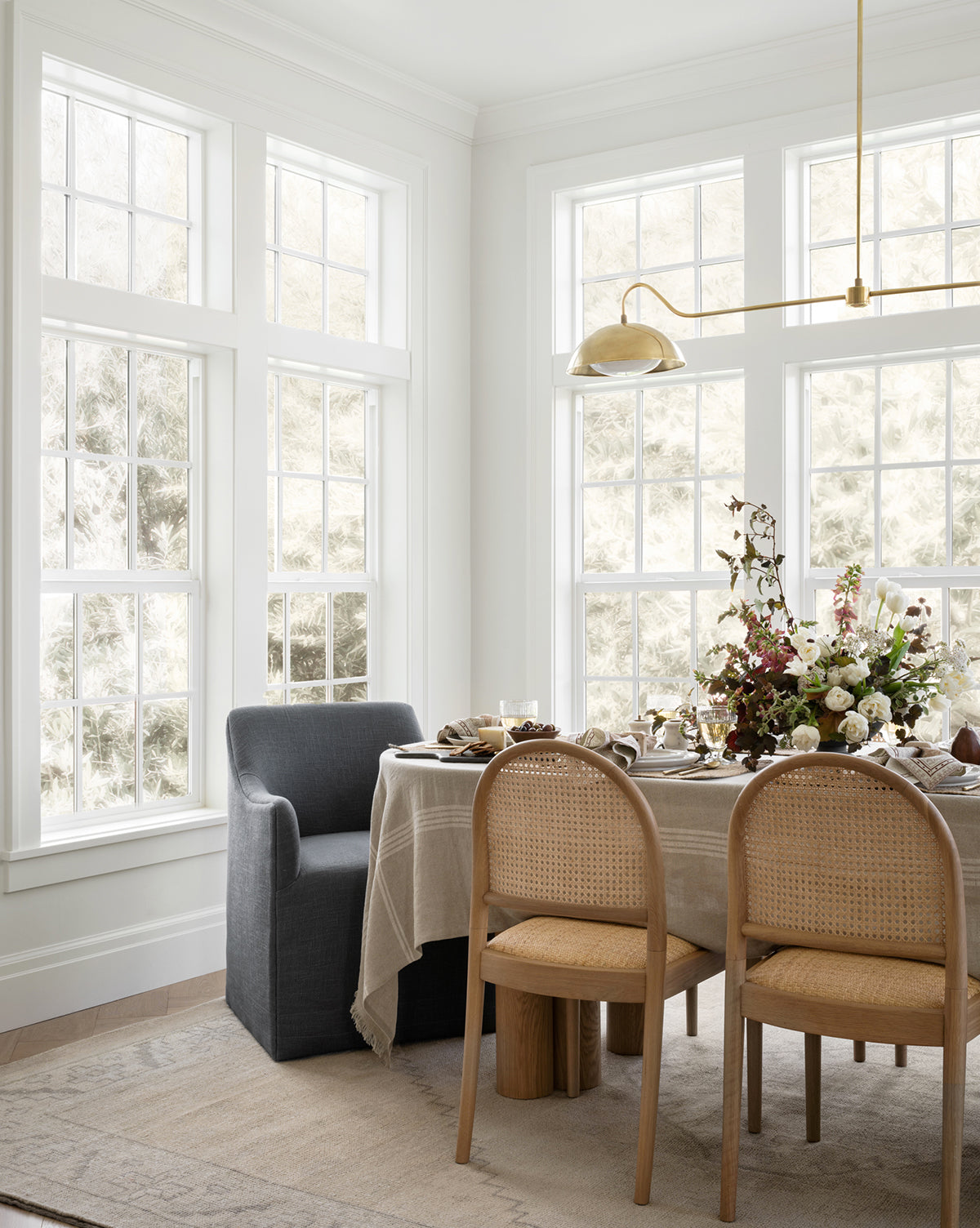 A bright dining area with large windows, a beige tablecloth, four chairs, and a floral centerpiece under a gold light fixture sits atop the Pembroke Hand-Knotted Wool Rug for vintage-inspired charm, by McGee & Co.