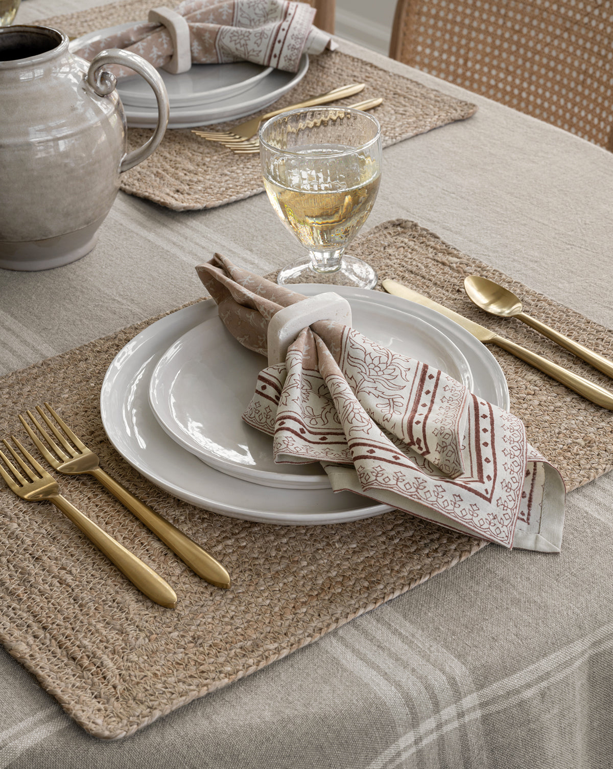 A close-up of a table setting with a woven placemat, Aiden Side Plate (Set of 4), patterned cloth napkin in a ring, gold cutlery, water glass with a light beverage, and beige ceramic pitcher on a neutral tablecloth, by McGee & Co.