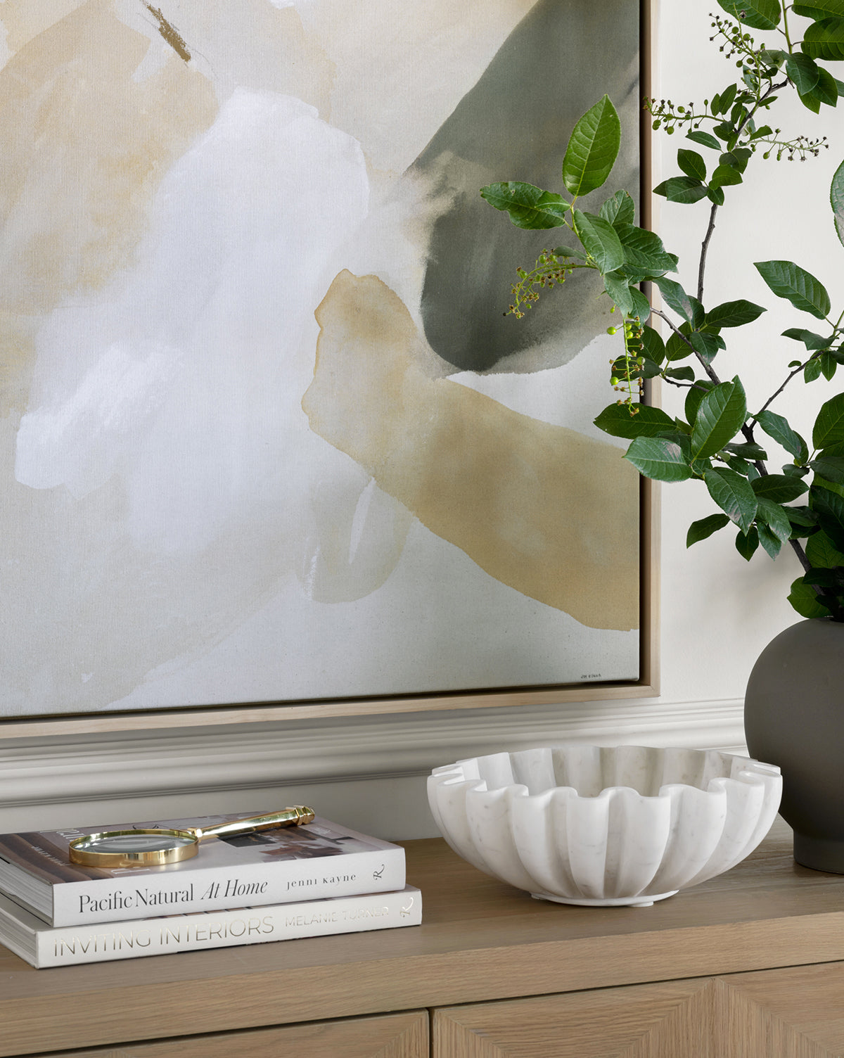 A modern abstract painting hangs above a wooden console table displaying decorative books, a gold magnifying glass, the Fluted Marble Bowl, and a gray vase filled with leafy green branches.