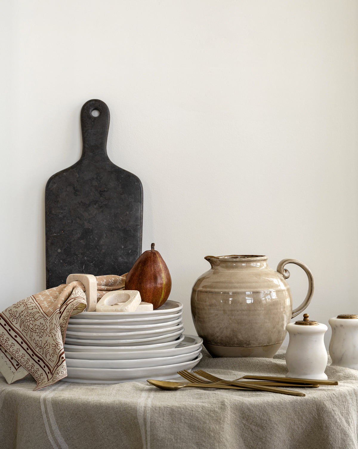 A stack of white plates with a napkin, bread, a pear, salt and pepper shakers, gold forks, and the Brown Stoneware Pitcher sits on a linen table beside a dark cutting board propped against the wall by McGee & Co.
