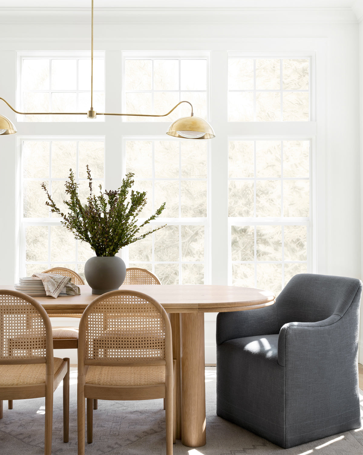 A bright dining room with large windows, a wooden table, and Elton Dining Chairs creates an inviting space. Add a gray linen blend armchair, modern gold light fixture, and a vase of green branches to complete the look, by McGee & Co.