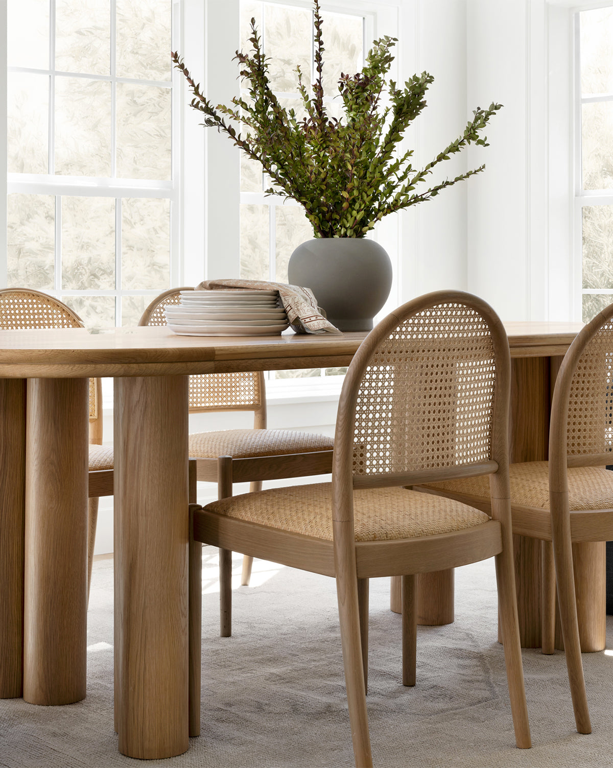 A light wood dining table with four Hadden Natural Dining Chairs featuring cane backs sits in a bright room. A gray vase with green branches and a stack of plates with napkins decorate the table near large windows, by McGee & Co.