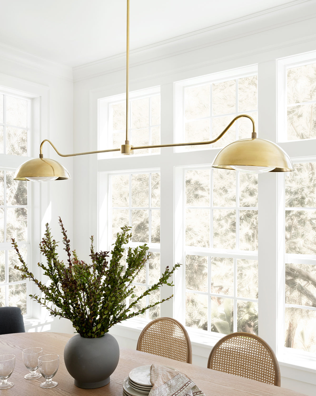 A modern dining area with a light wood table, rattan chairs, a black vase with greenery, and the Aldorno Linear Chandelier by Makerspalm, set before large sunlit windows that frame a snowy outdoor view. McGee & Co.