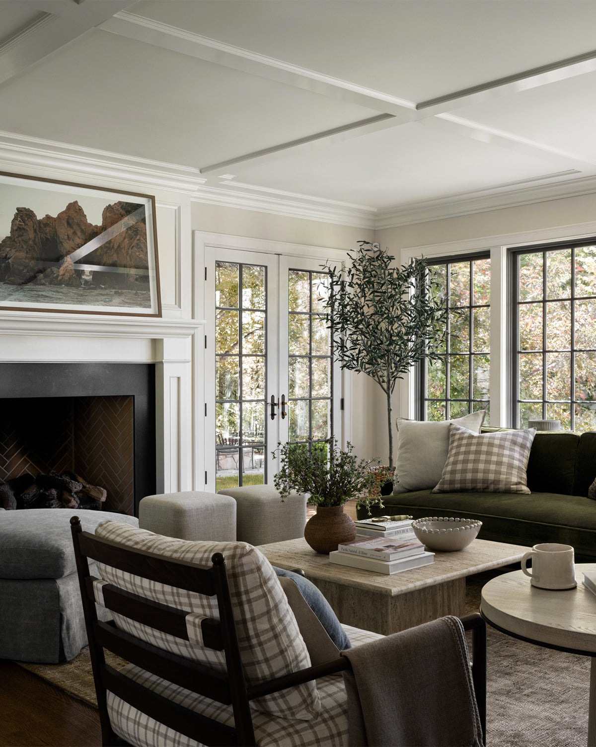 A bright, elegant living room features large windows, a green sofa with plaid cushions, a cozy armchair, and modern fireplace. The Faux 96 Olive Tree with Woven Pot brings warmth and natural charm to this inviting, sun-filled space.