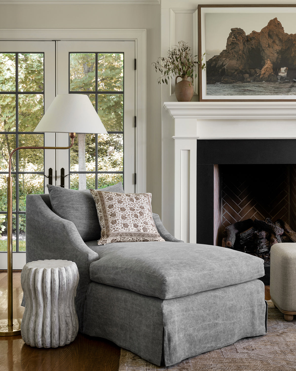 A cozy living room corner with a gray chaise lounge, patterned pillow, textured side table, Wainwright Swoop Floor Lamp, and fireplace. Large windows reveal greenery; a landscape photo hangs above the mantel, by McGee & Co.