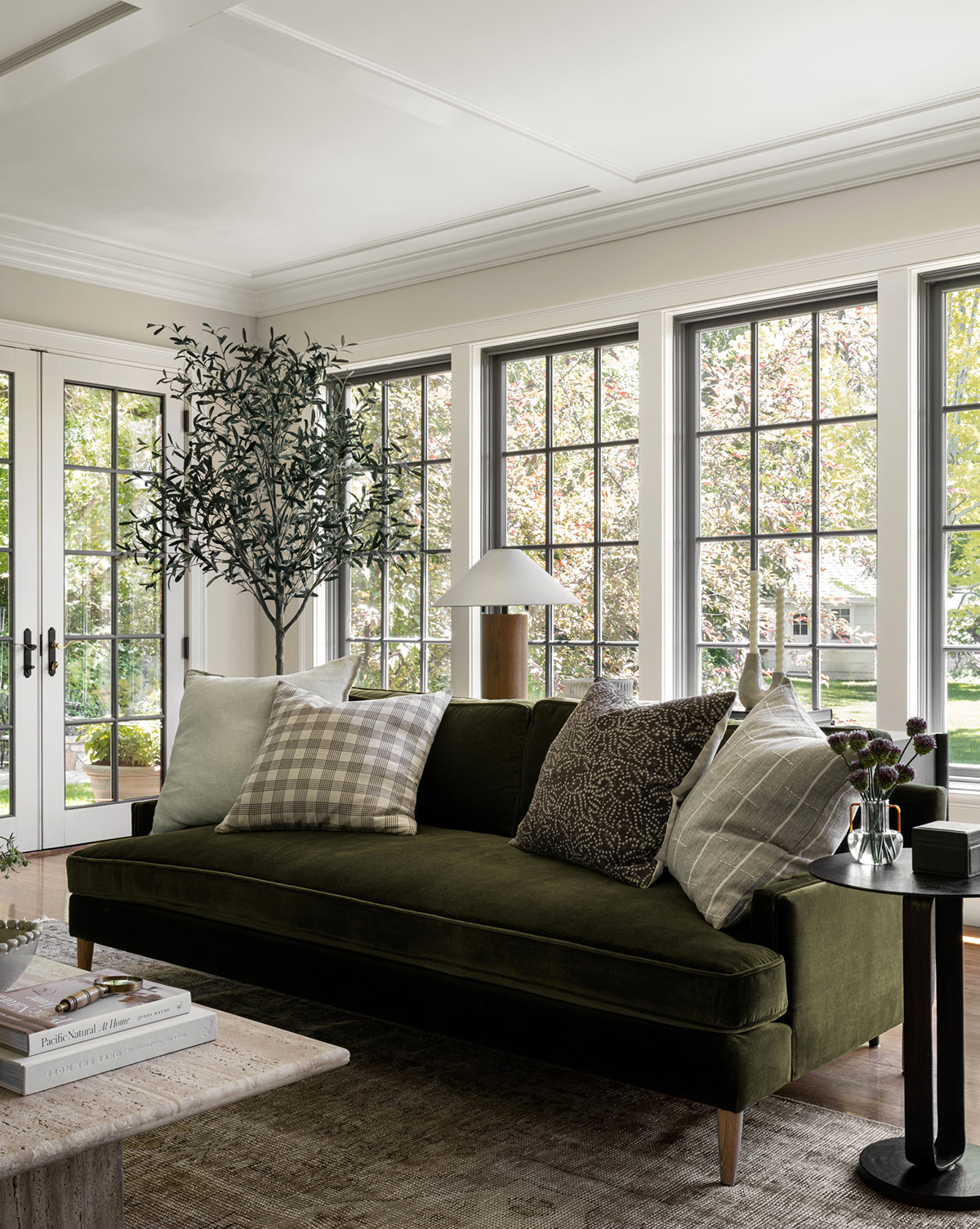 A modern living room with large windows, a green Makerspalm Evan Sofa 84 with patterned pillows, a side table and lamp, potted tree, and coffee table with books—all bathed in natural light. McGee & Co.