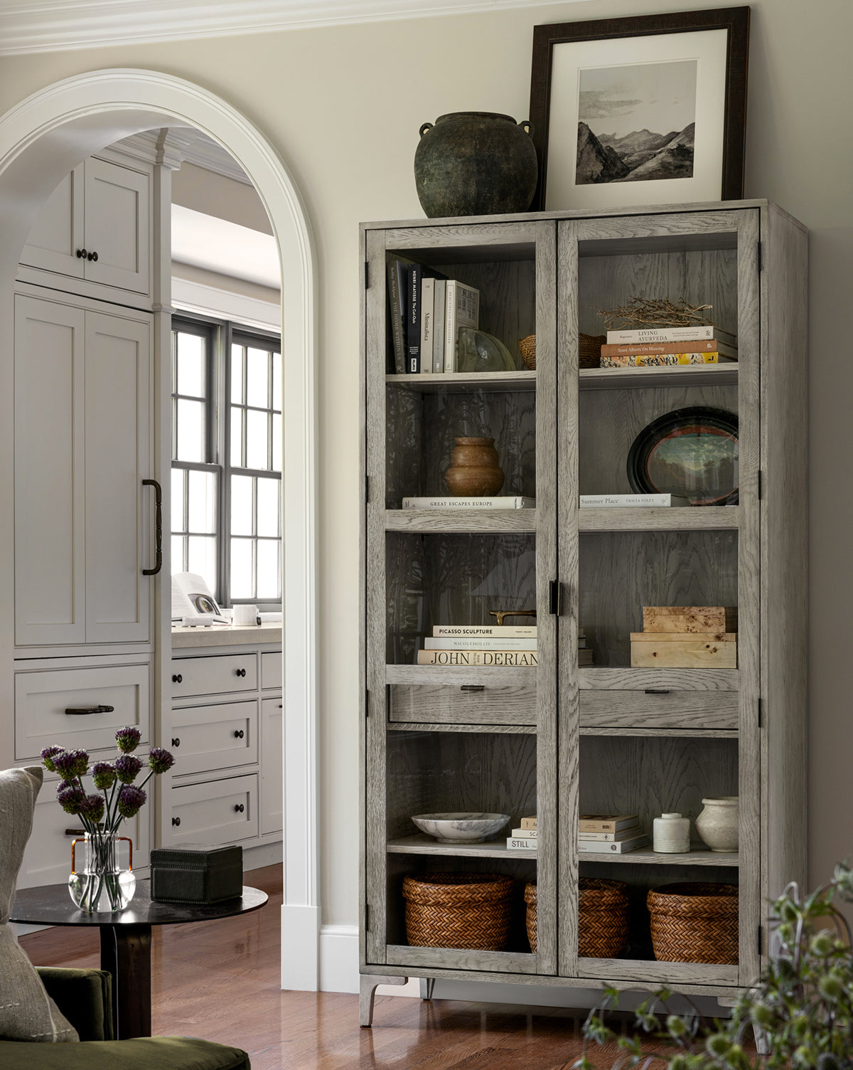 A tall, light wood cabinet with glass doors displays books, pottery, baskets, and the Monochrome Mountains print in a cozy living space. An archway reveals a kitchen with white cabinetry in the background.