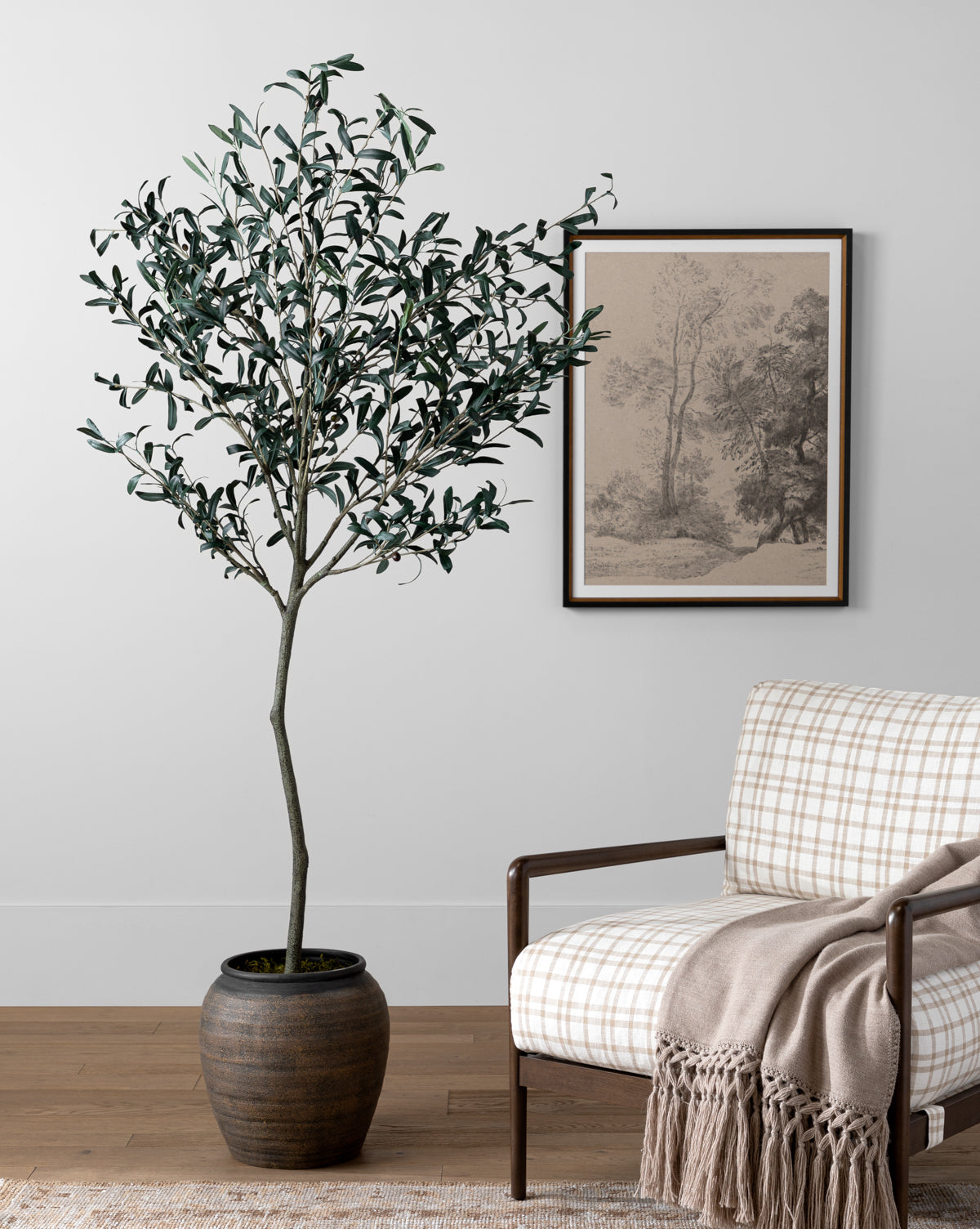 The Faux 72 Olive Tree with Ceramic Pot stands beside a plaid armchair and beige throw blanket. A framed black and white landscape drawing hangs on the light gray wall above the wooden floor, creating inviting indoor decor.
