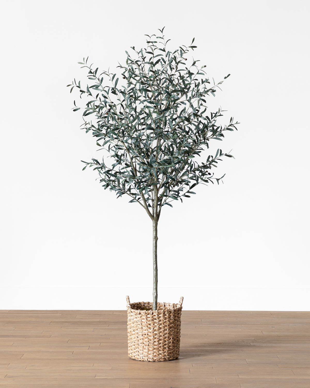 The Faux 96 Olive Tree with Woven Pot features slender green leaves and stands on a wooden floor, adding chic olive tree decor against a plain white background.