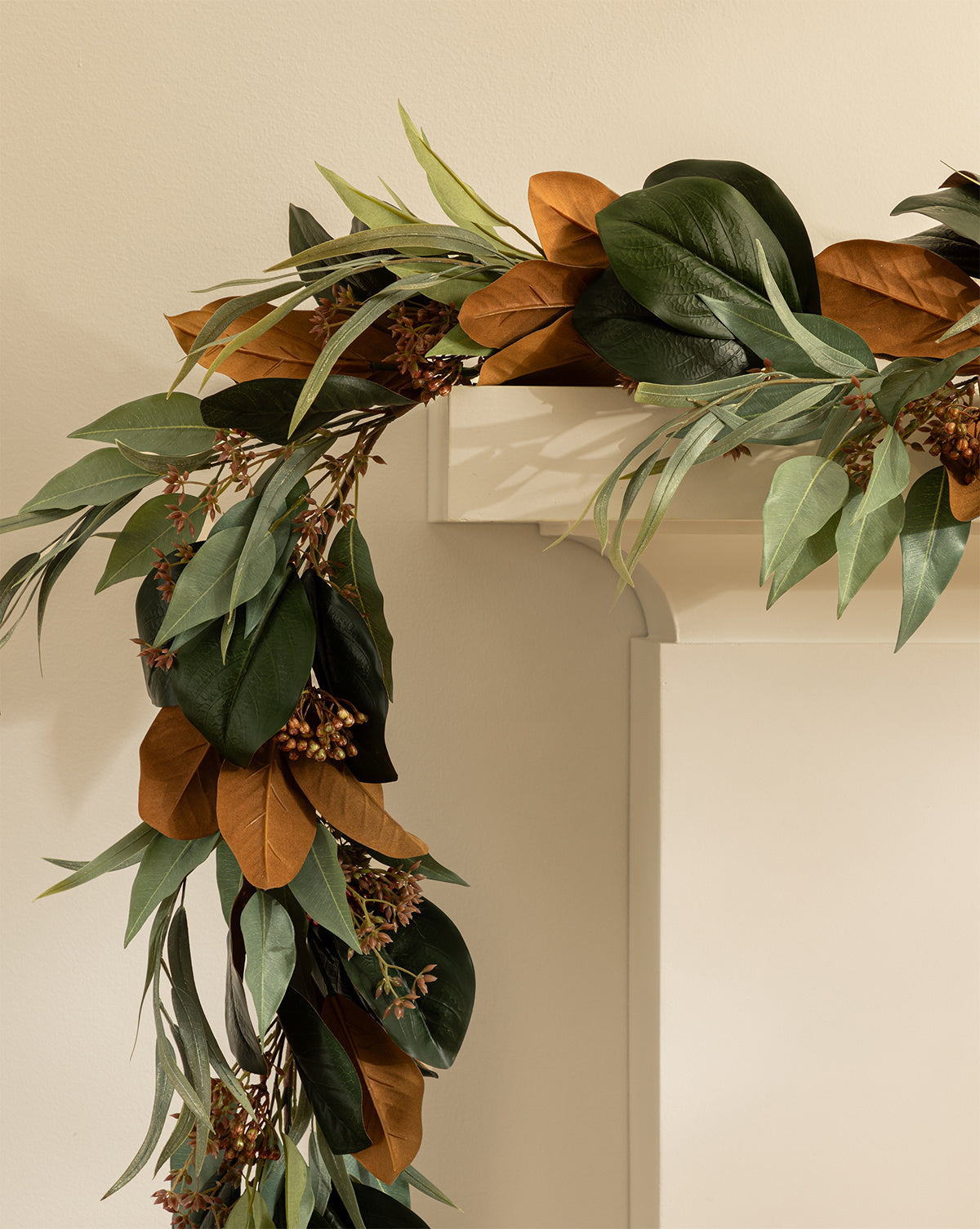 The Faux Magnolia & Eucalyptus Garland, featuring green and brown leaves with small berry clusters, drapes over a white mantel, creating an ideal winter accent against the beige wall, by McGee & Co.