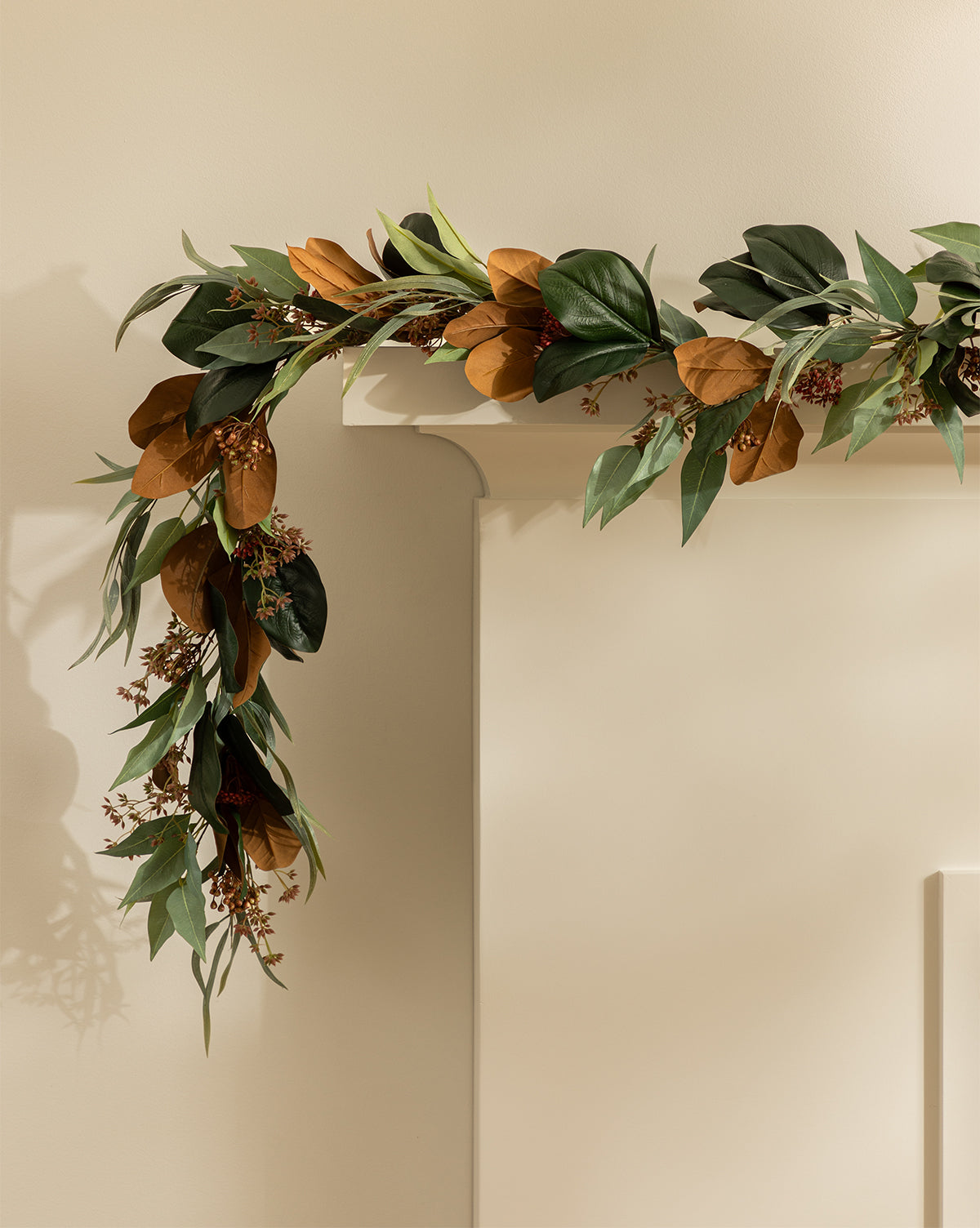 The Faux Magnolia & Eucalyptus Garland, featuring green and brown leaves, drapes over a cream mantel against a light beige wall, offering an elegant winter accent for your décor, by McGee & Co.