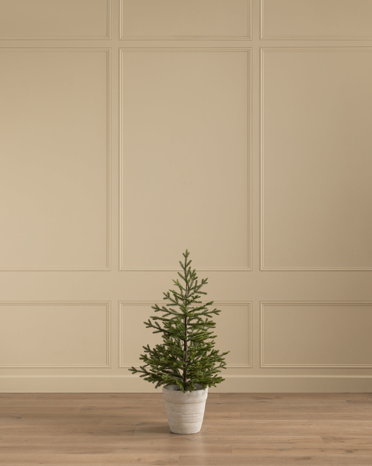 The Faux Spruce Pre-Lit Potted Tree is displayed on a wooden floor by a beige paneled wall, adding a minimalist, neutral accent to the room, by McGee & Co.
