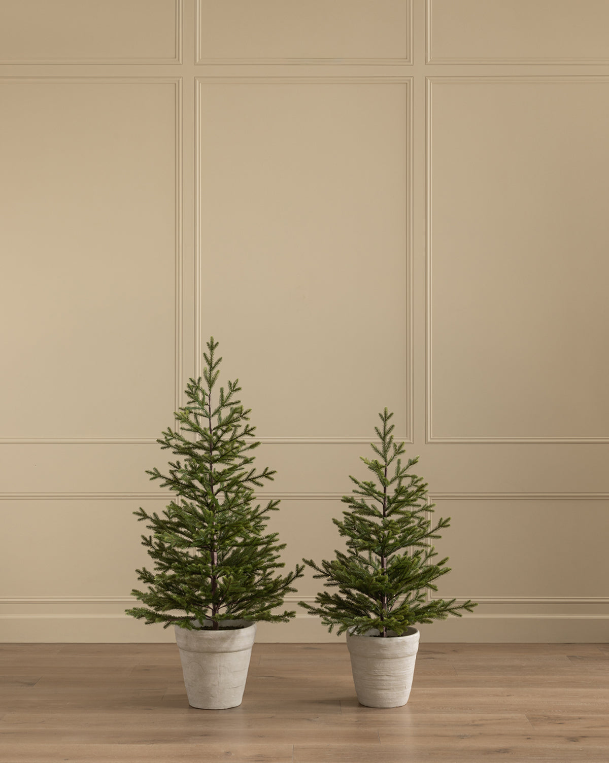 A Faux Spruce Pre-Lit Potted Tree stands on a light wooden floor beside another potted faux tree of different height, set against a beige paneled wall, by McGee & Co.