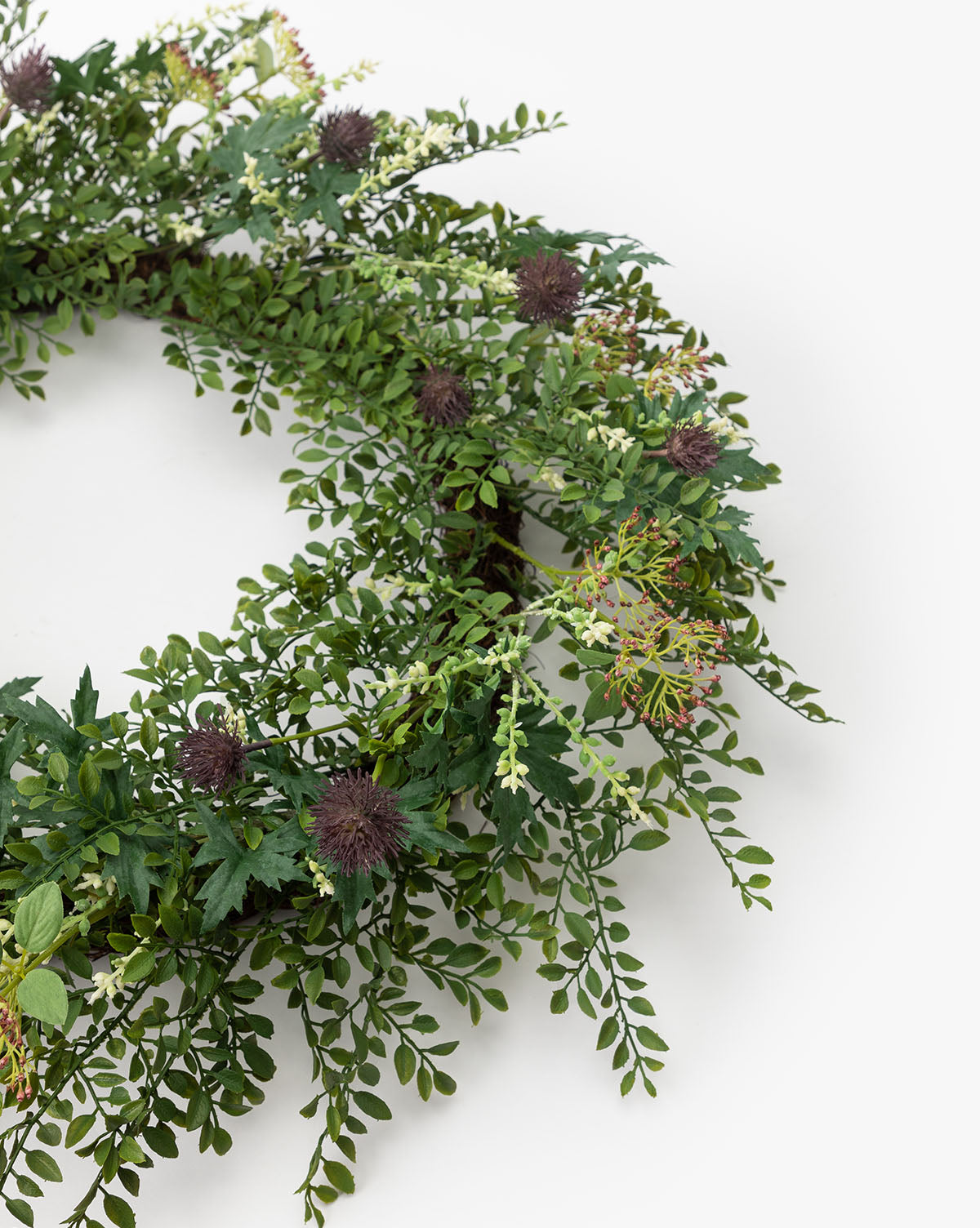 The Faux Summerset Wreath features assorted faux greenery and small, dark purple thistle-like flowers in a circular design—ideal for seasonal decor and displayed against a plain white background.