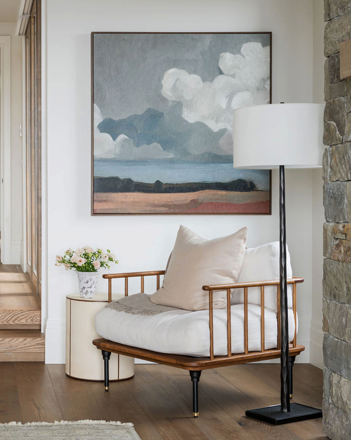 A cozy reading nook features a cushioned wooden chair, large pillow, round side table with a flower vase, tall floor lamp, and Feathered Clouds—an abstract landscape painting—hanging on a white wall near a stone accent.