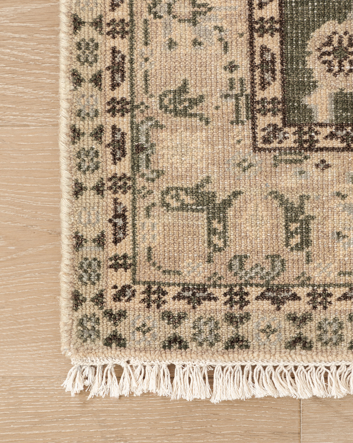 A close-up of the Fernwood Hand-Knotted Wool Rug shows its intricate beige, green, and pink motifs on a light wooden floor, with delicate white fringes lining the edge, by McGee & Co.