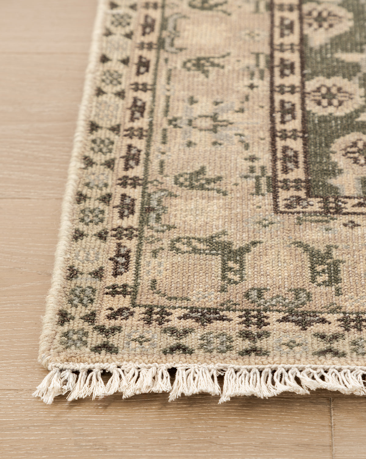 A close-up of the Fernwood Hand-Knotted Wool Rug shows its beige and green wool, fringed edges, and geometric floral motifs, elegantly placed on a light wooden floor, by McGee & Co.