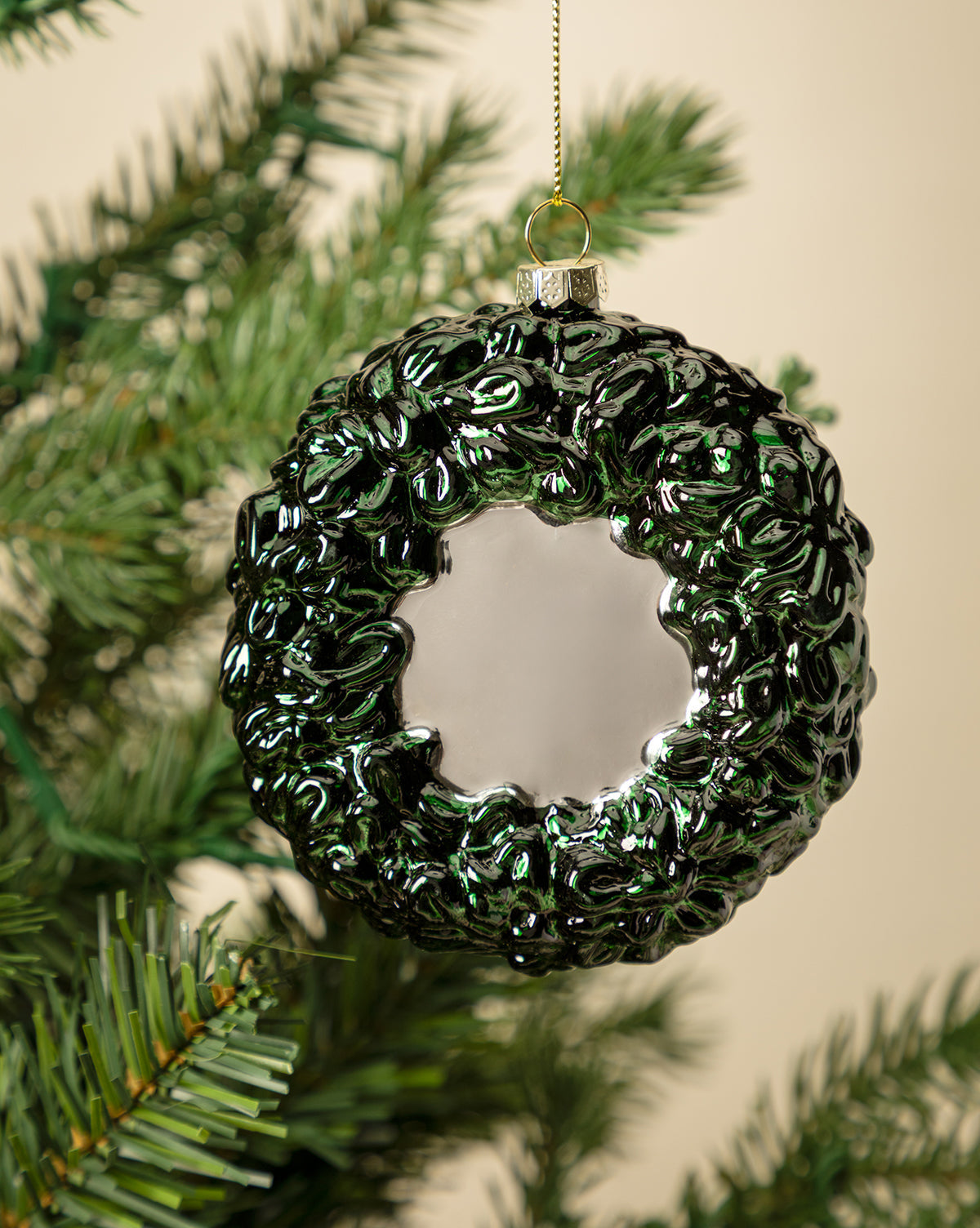 The Festive Wreath Ornament, dark green and shiny with a circular hollow center, hangs elegantly on a Christmas tree branch—an ideal 2025 keepsake set against a softly blurred, neutral background, by McGee & Co.