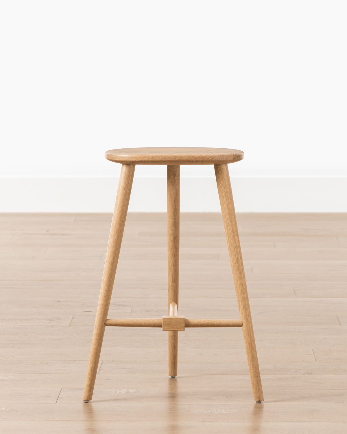 The Fielder Counter Stool features a round seat and three legs in a light oak finish, shown on a light wood floor beside a white wall, by McGee & Co.