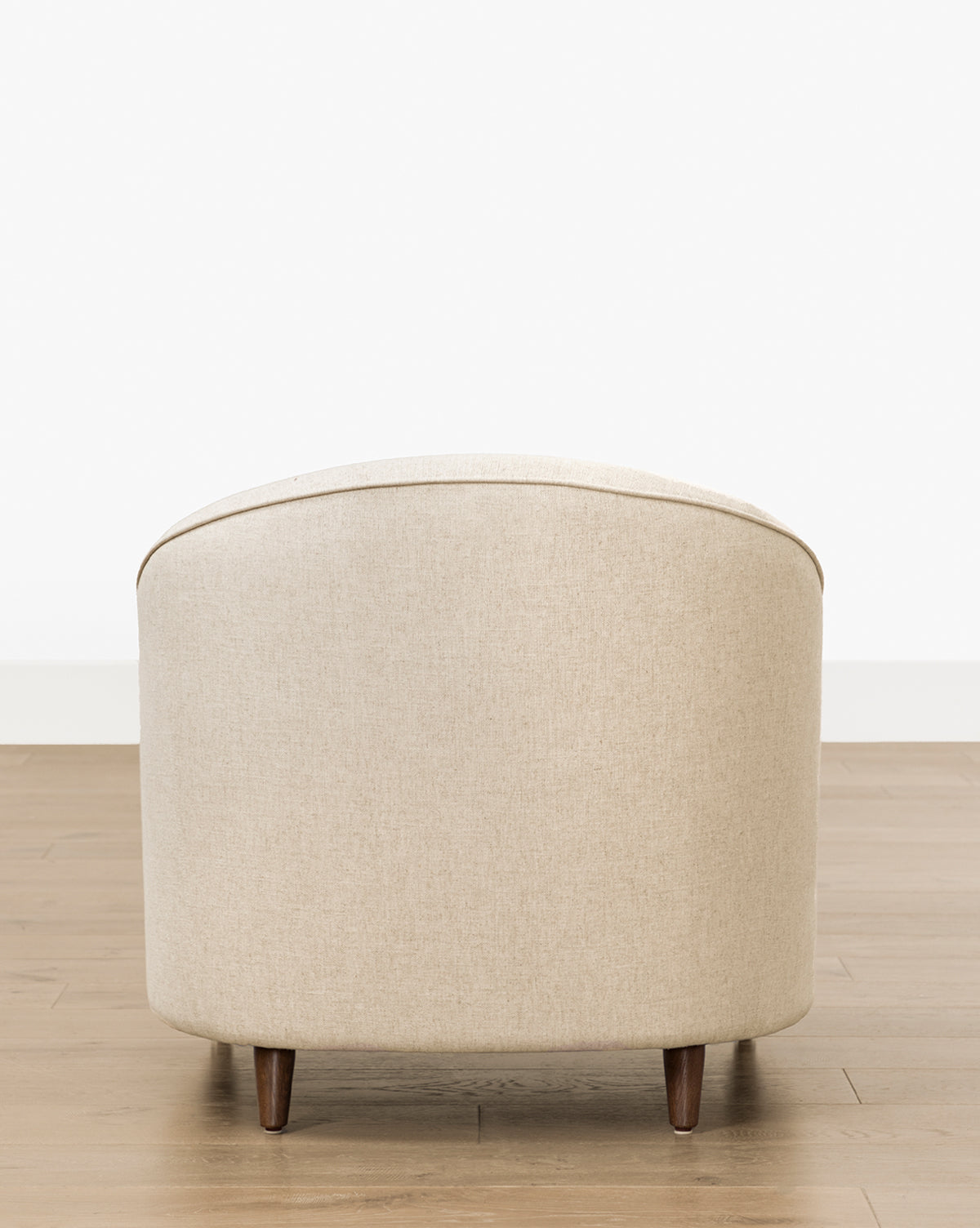 The Fletcher Lounge Chair (Ready to Ship) is a beige upholstered armchair with a rounded back and dark wooden legs, set on a light wood floor against a plain white wall and viewed from the back by McGee & Co.