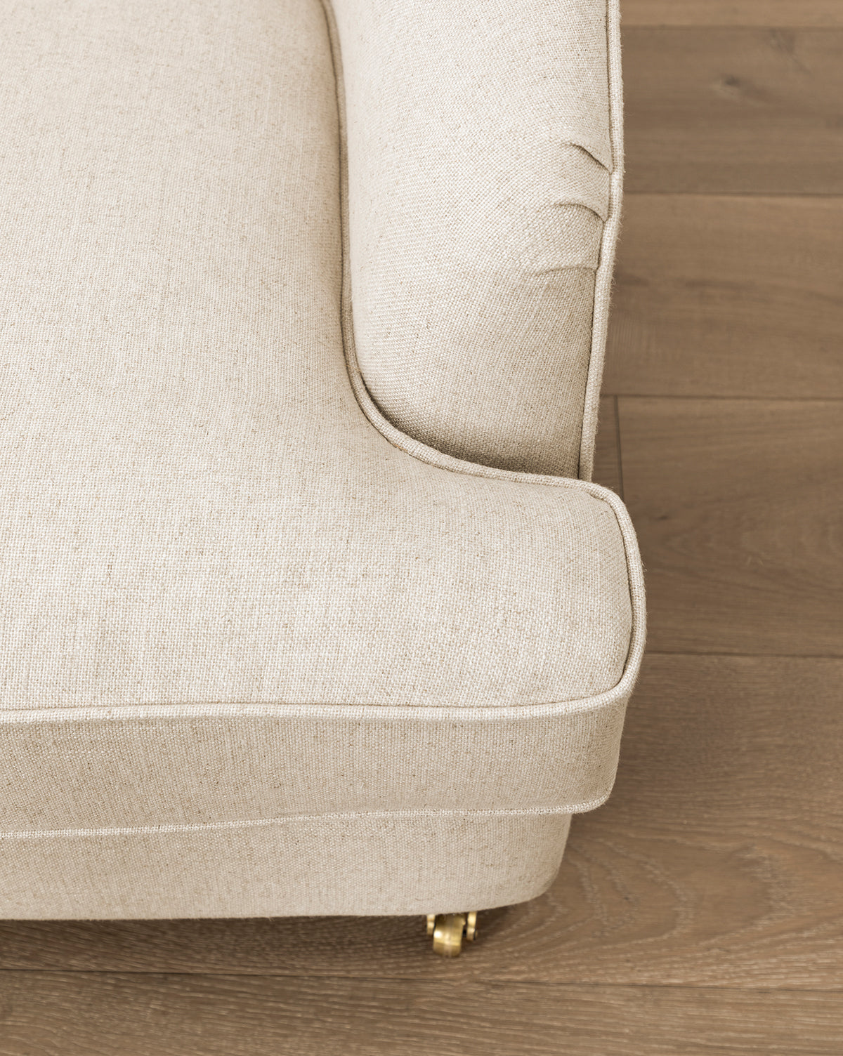Close-up of the corner of the Fletcher Lounge Chair (Ready to Ship) in beige, featuring piped edges and a visible brass wheel beneath the leg, set on a wooden floor by McGee & Co.