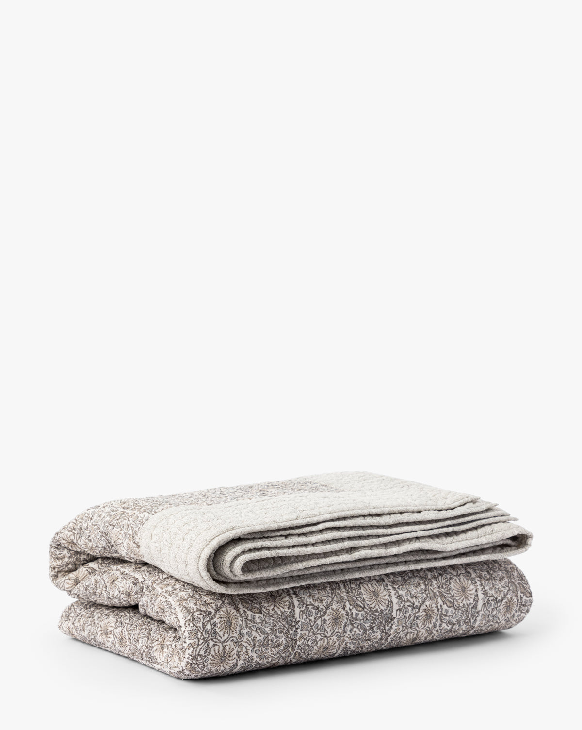 A Floral Block Print Quilt in neutral tones, folded to display its pattern, rests atop a hand-quilted textured blanket, both set against a plain white background.