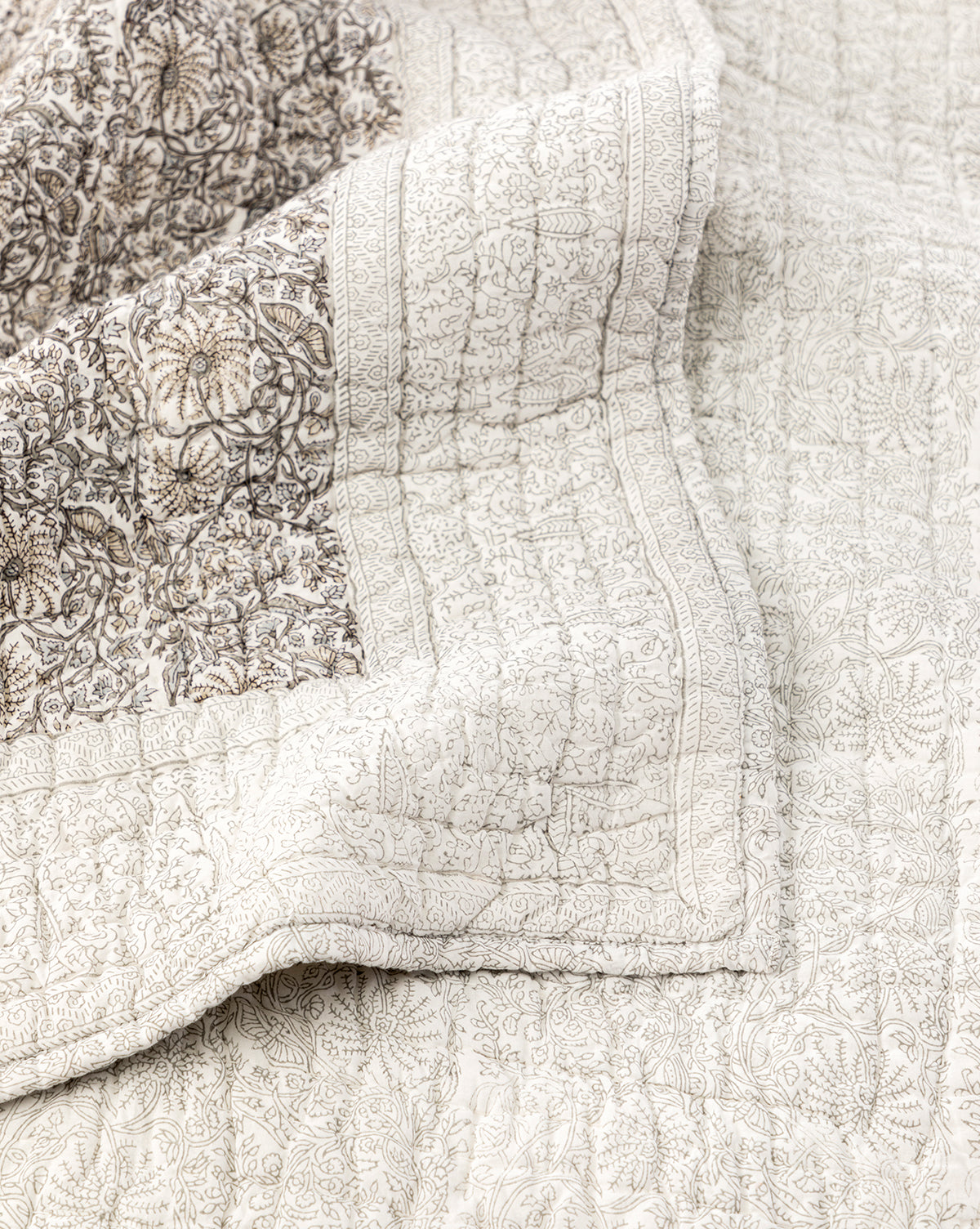 A close-up of the Floral Block Print Quilt highlights its cream color, beige and brown floral and paisley patterns, a folded corner revealing detailed hand-quilted stitching and the elegant block printed bedding design.