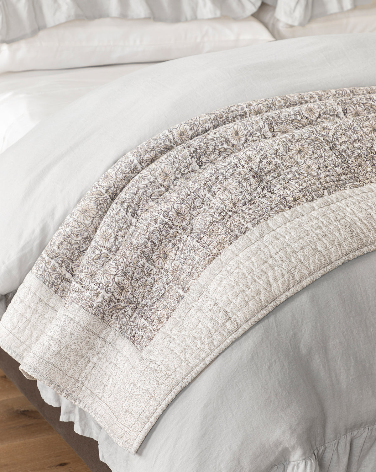 A close-up of a bed with the Floral Block Print Quilt, showcasing its light-colored floral pattern bordered by solid fabric, elegantly draped over crisp white bedding.